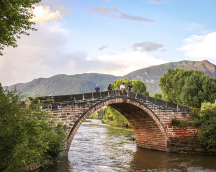 A stone arch bridge over a river surrounded by trees, with mountains and a partly cloudy sky in the background, and people walking across the bridge.