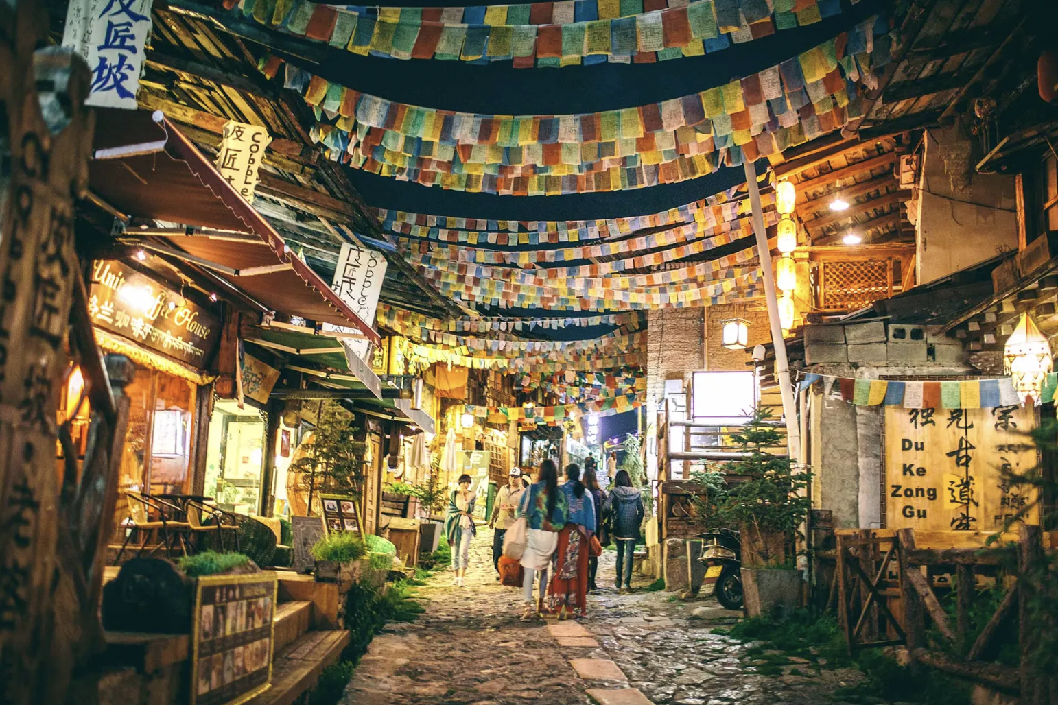 Night scene of a lively street with hanging colorful prayer flags overhead, small shops and cafes lining the cobblestone street, and people walking and socializing.