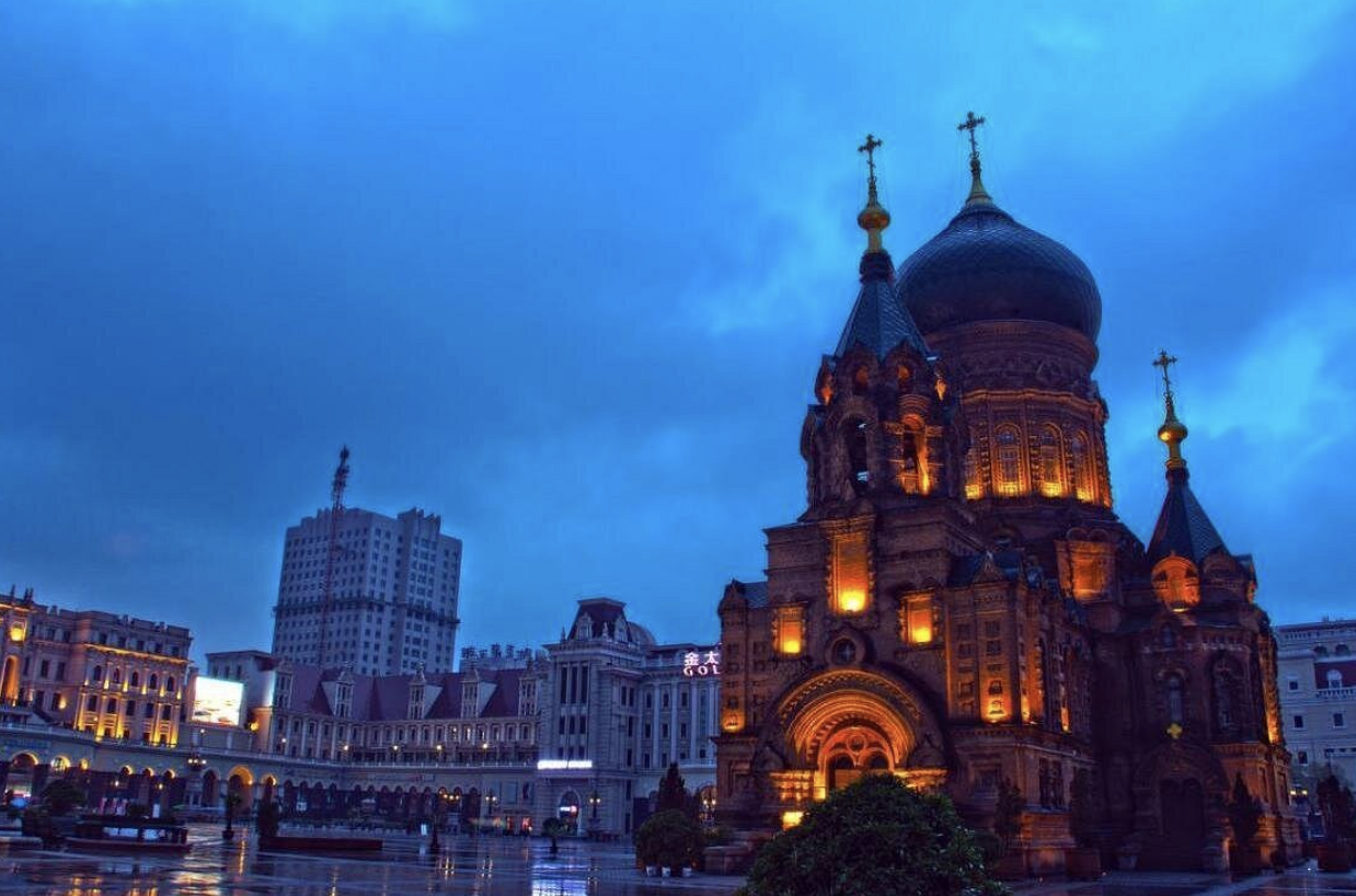 A large illuminated church with onion domes and crosses on top, set against a blue sky with some clouds, surrounded by buildings and a wet plaza.