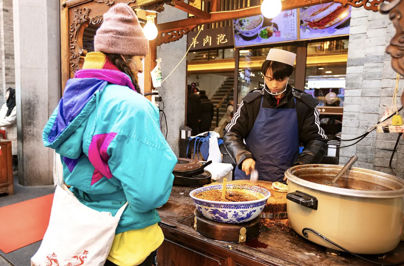 A woman in colorful winter clothes and a hat stands at a food stall, ordering spicy soup from a young man chef who is preparing food. The stall is decorated with traditional wooden carvings, and there are large bowls and a slow cooker on the counter.