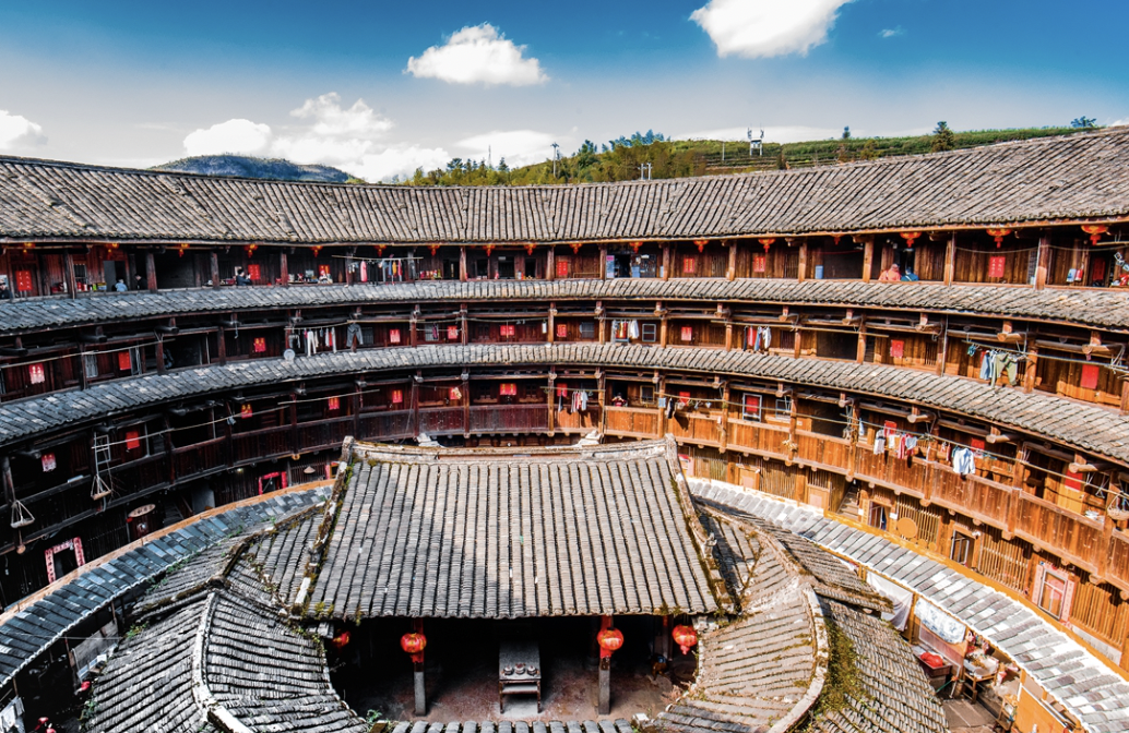 Traditional circular multi-story wooden building with balconies, red lanterns, and laundry hanging, situated in a rural area with hills in the background.