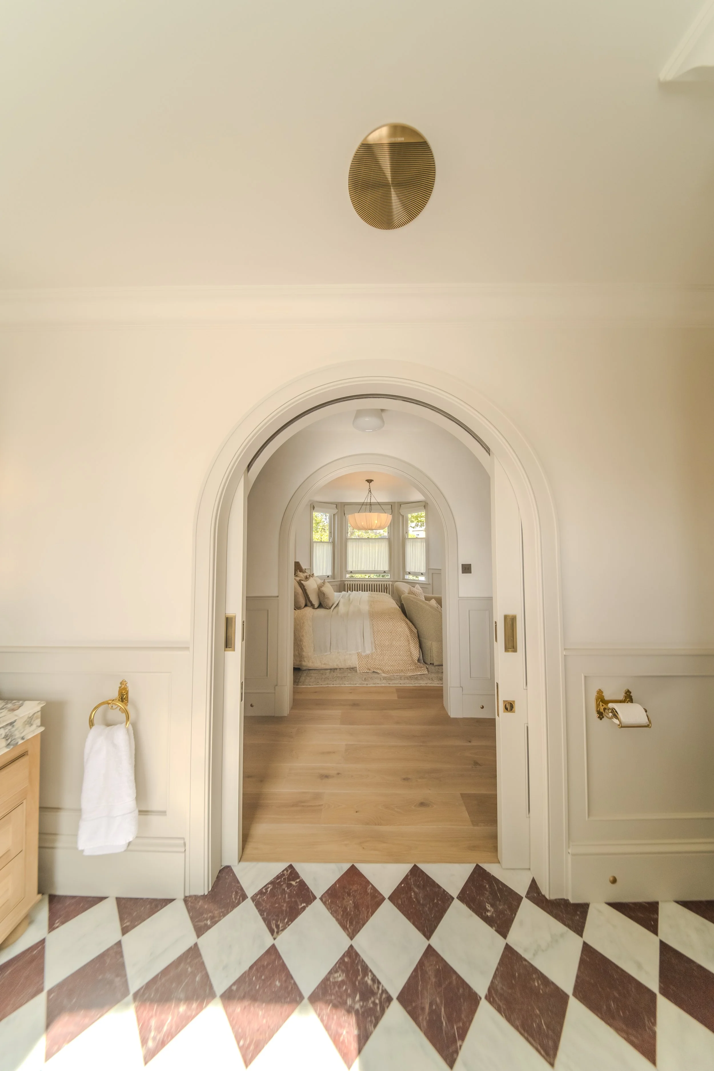 Jamie and Sophie Laing arched bathroom entrance. Interior view of a bedroom seen through an arched doorway, with a bed, windows, and a chandelier in the background, and a marble and tile floor in the foreground.