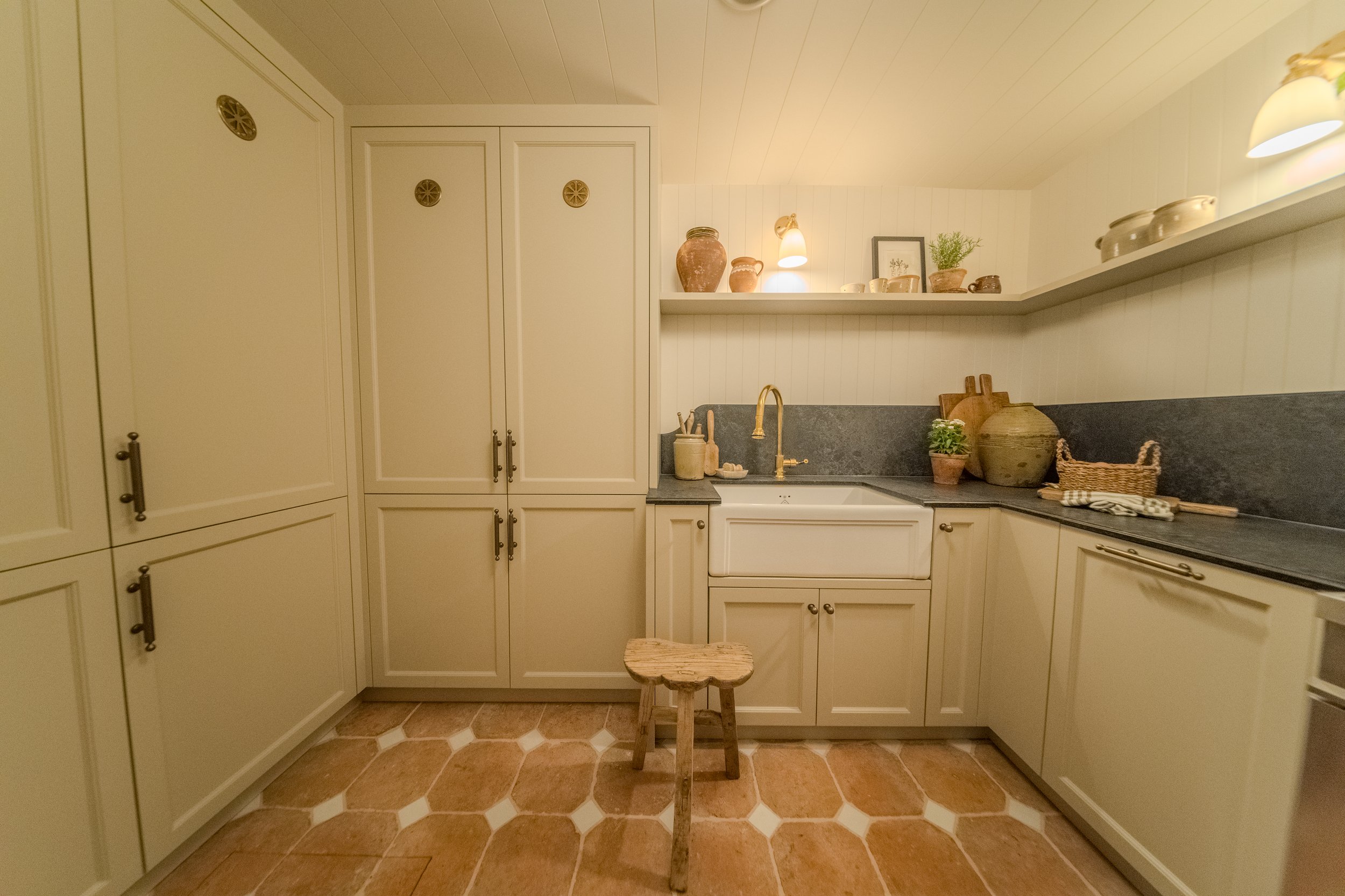 A cozy kitchen with cream-colored cabinets, a white farmhouse sink with a brass faucet, black countertop, and decorative pottery on shelves. The kitchen has warm lighting, a terracotta tiled floor, and a small wooden stool in front of the sink.