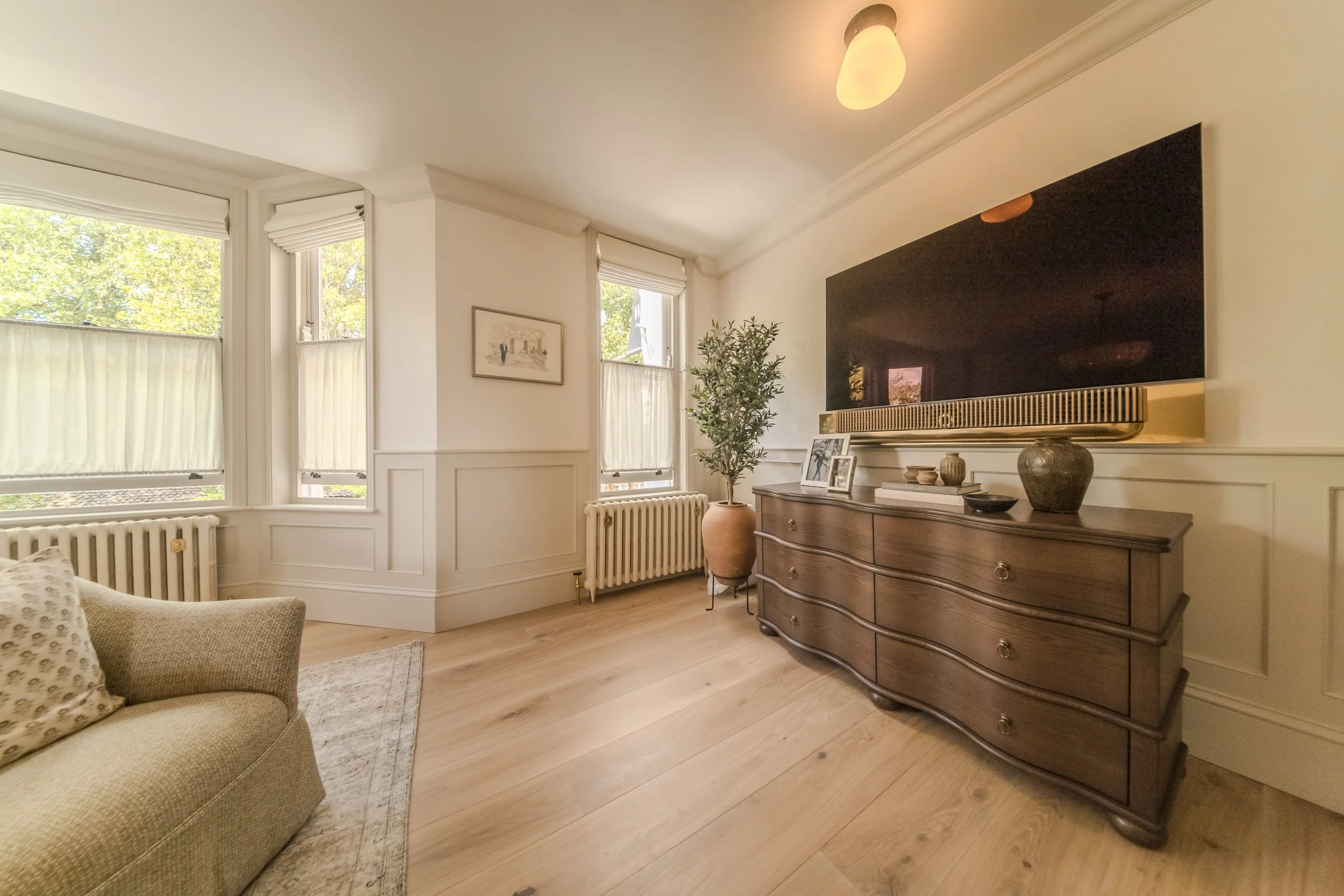 Jamie and Sophie Laing new home bedroom room with beige sofa, wooden dresser, flat-screen Bang & Olufsen TV, potted plant, and windows with white blinds