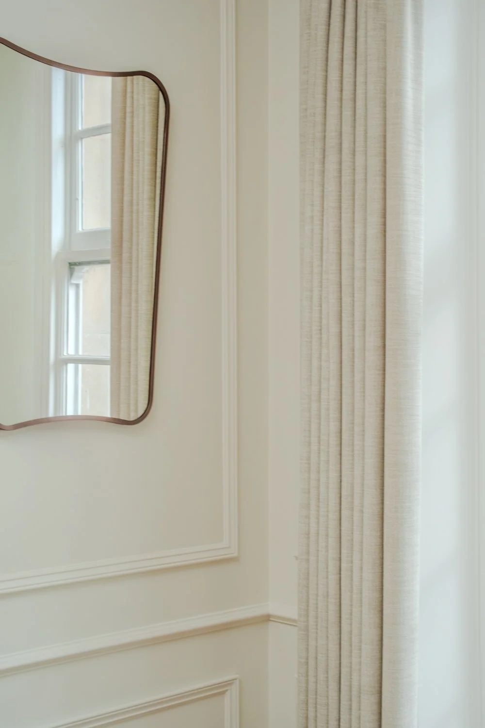 A room corner with a wavy-edged mirror on the wall and a window with beige curtains.