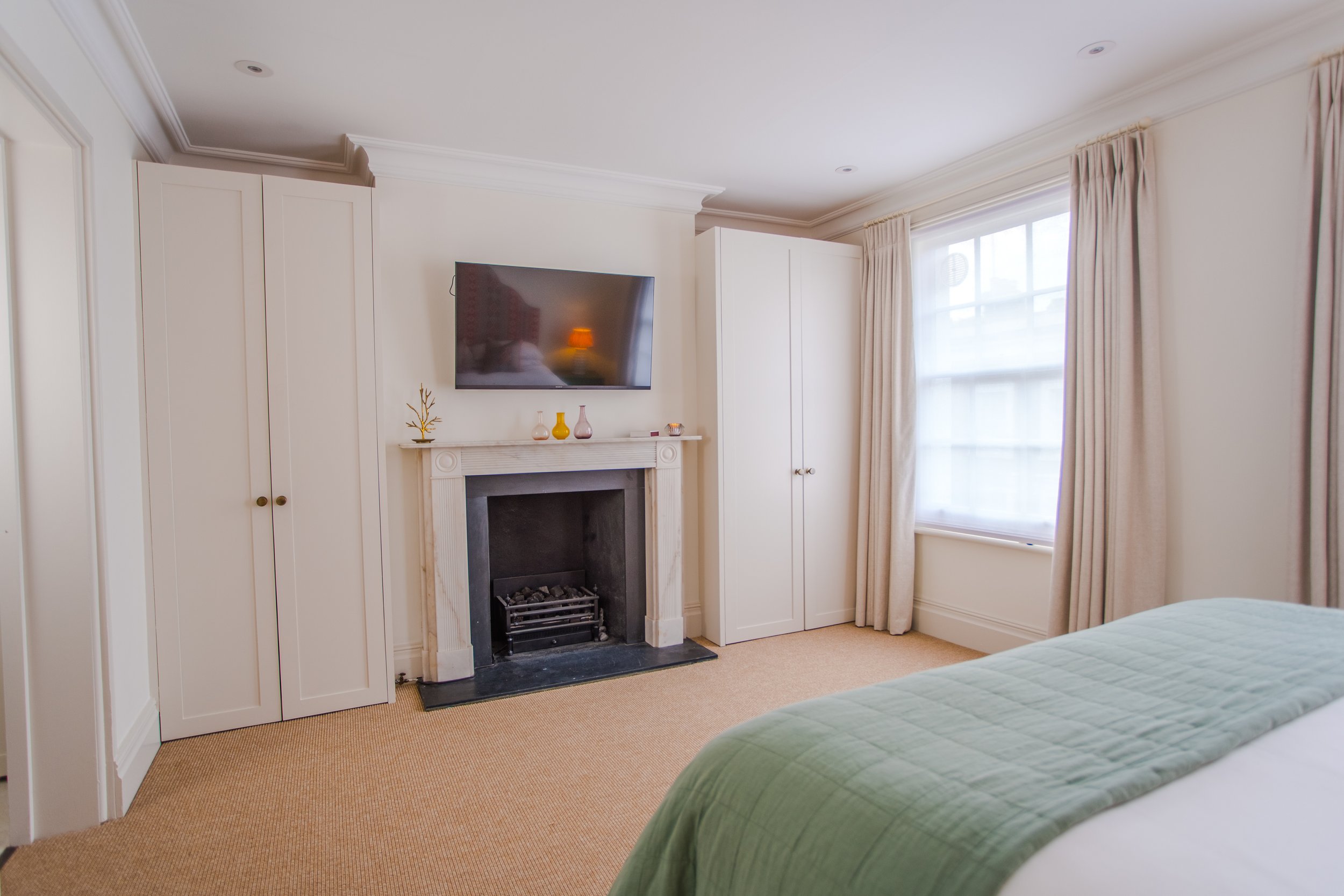 Hotel room with a bed covered in a green bedspread, a window with beige curtains, a fireplace with decor and a wall-mounted TV above, off-white walls, and carpeting.