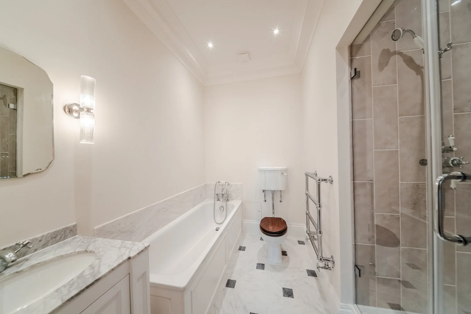 A clean, modern bathroom in Mayfair with a bathtub, a toilet with a wooden seat, a glass shower enclosure, a marble sink countertop, and white tiled walls and floor. Designed by Kensington Design Ltd 