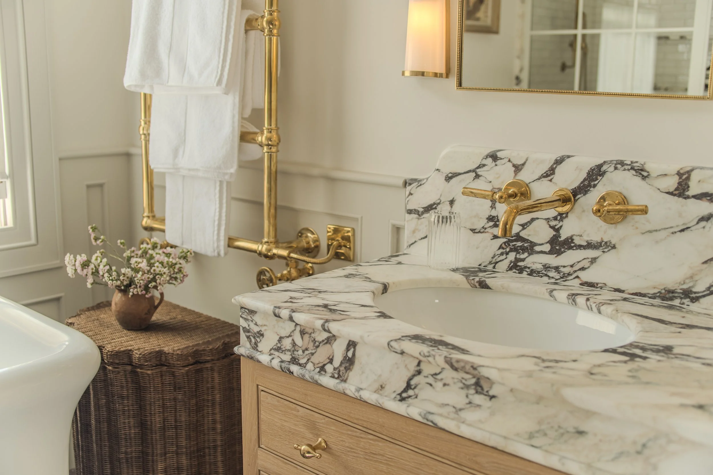 Jamie and Sophie Laing bathroom with marble sink, gold fixtures, towel rack, and a wicker table with a vase of pink flowers. Desinged by House 9 design 