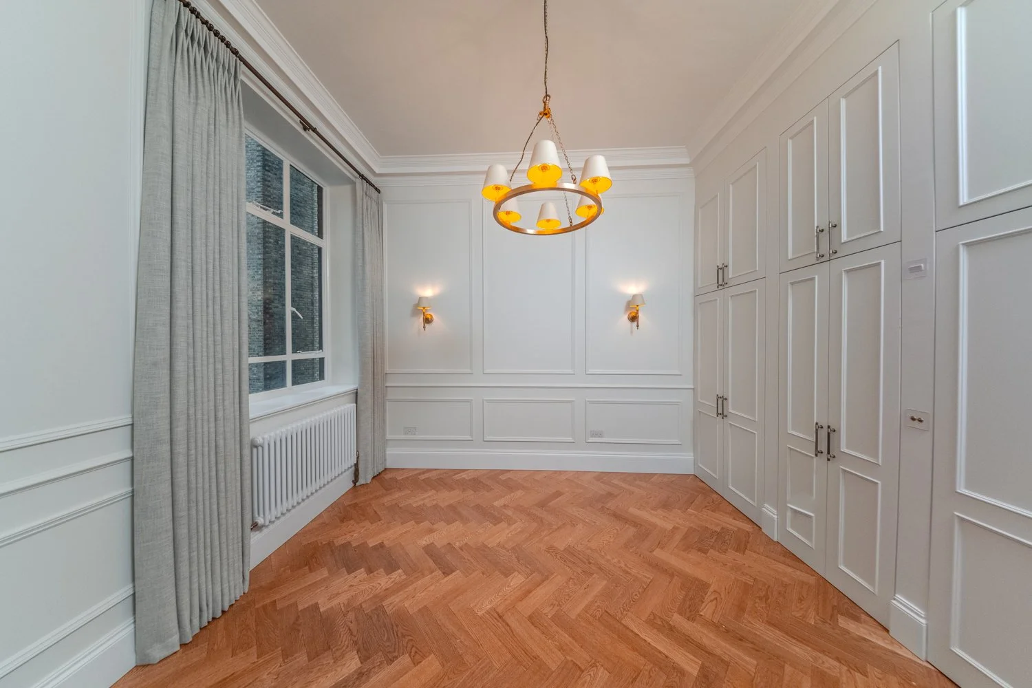 Empty room with white paneled walls, hardwood herringbone floor, large window with white curtains, and built-in white cabinets. There is a decorative chandelier hanging from the ceiling and two wall sconces on either side of the window. 