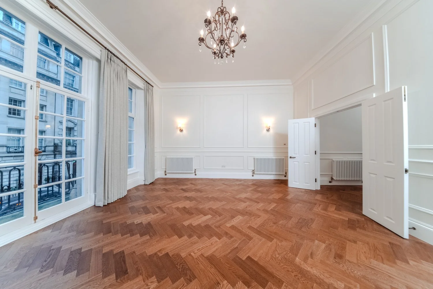Empty room with large windows, white walls, hardwood floor, chandelier, and wall-mounted lights.