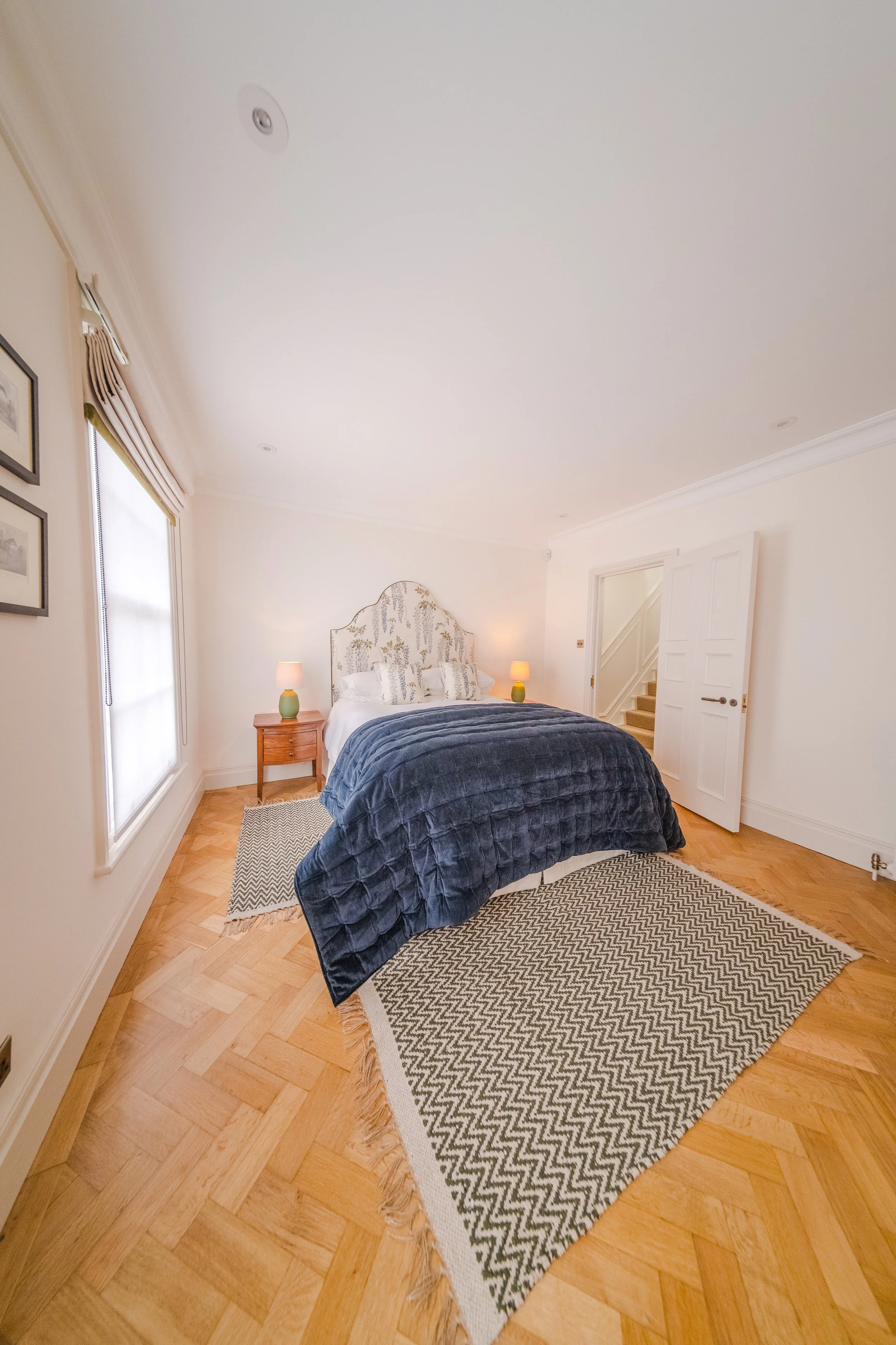 A bedroom with a large bed covered in a dark quilt, two side tables with lamps, a patterned headboard, and a zigzag rug on a hardwood floor. There are windows with curtains and framed pictures on the wall.