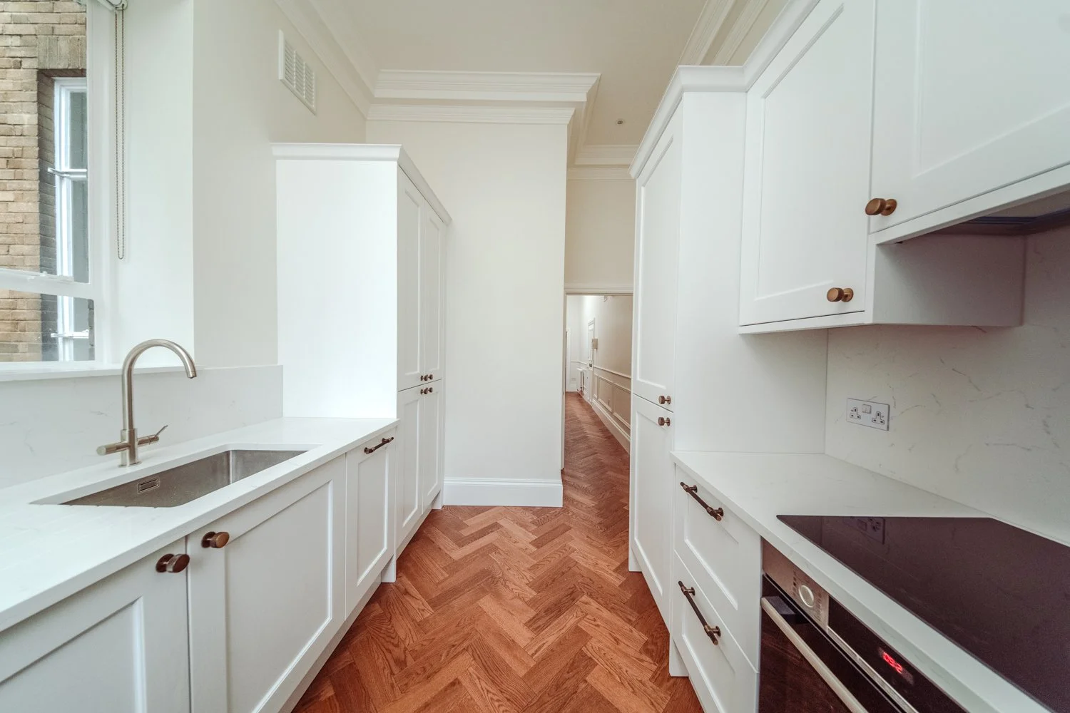Mayfair modern kitchen with white cabinets, wooden flooring, and a window with a view of brick building exterior. Designed by Kensington Design Ltd 