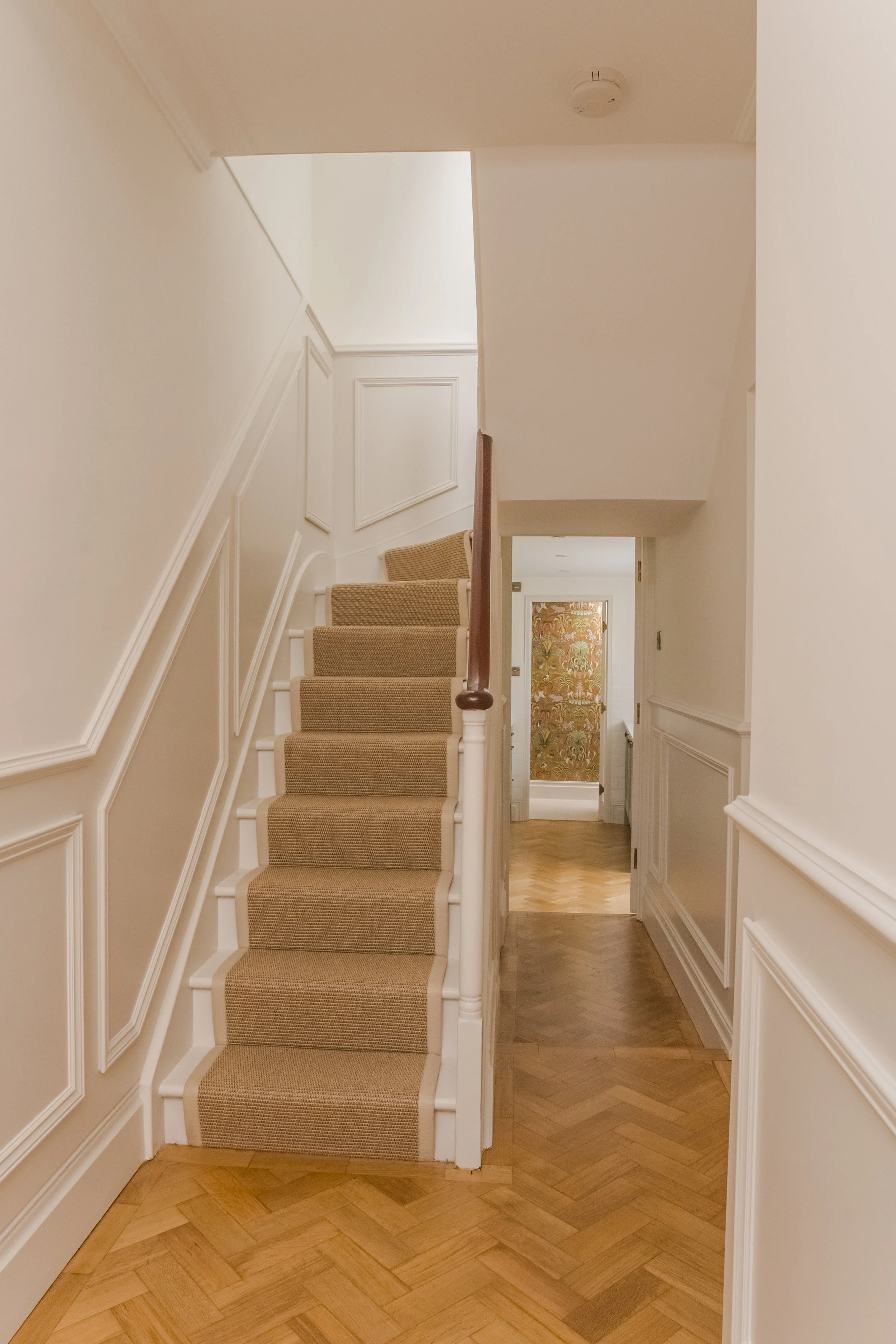 Interior view of a hallway with wooden flooring, a staircase with beige carpet, and white paneled walls leading to upper floors and rooms.