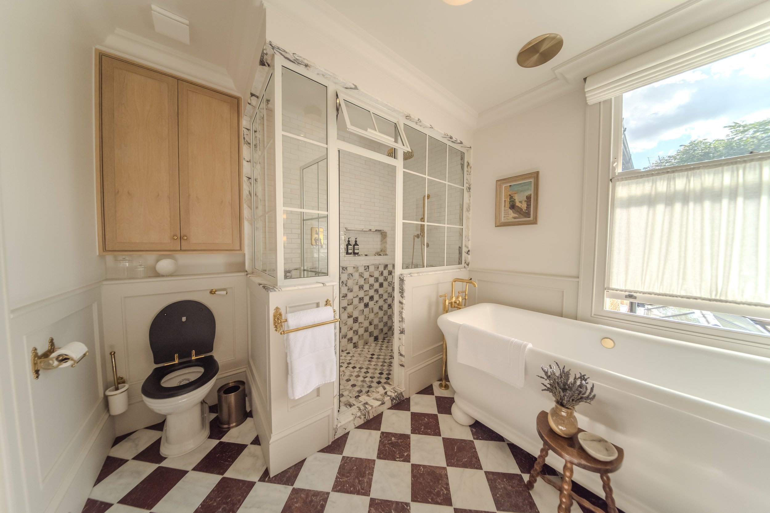 Jamie and Sophie Laing renovated vintage-style bathroom with a clawfoot bathtub, a separate shower with glass windows, a toilet with a black seat, wooden cabinets, a framed painting, and patterned marble flooring. Designed by House 9 designers. 