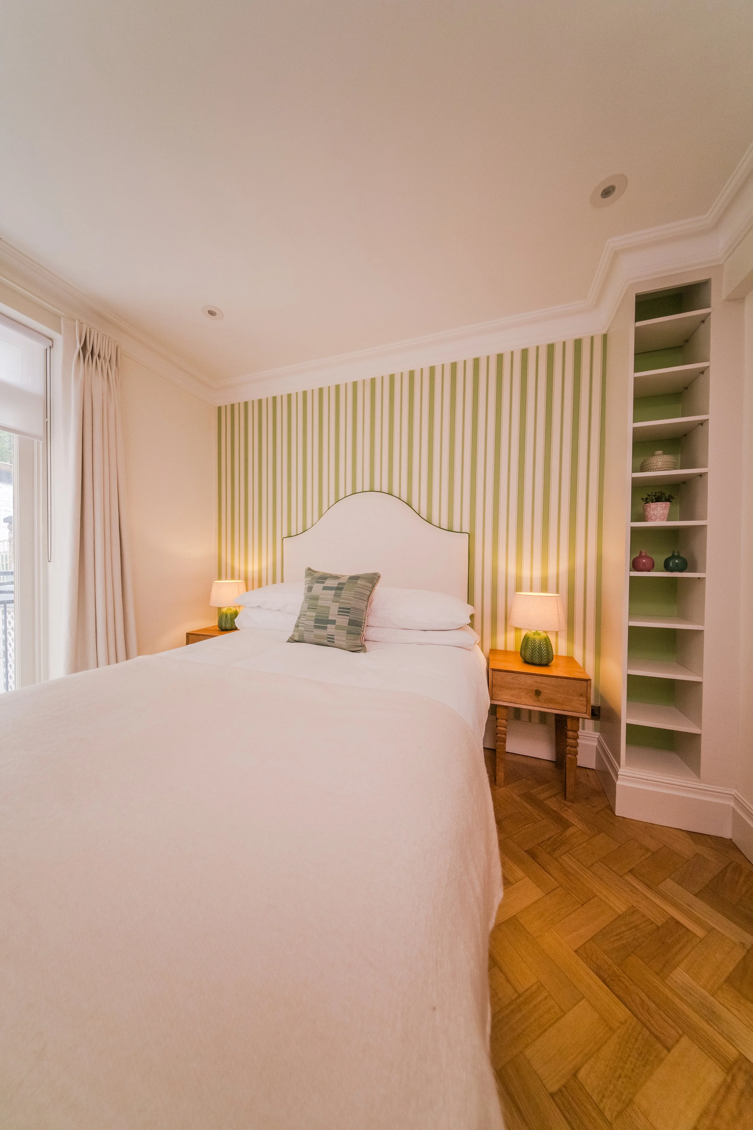 Cozy bedroom with a white bed, striped green and white accent wall, two bedside tables with lamps, and built-in shelving on the right side.