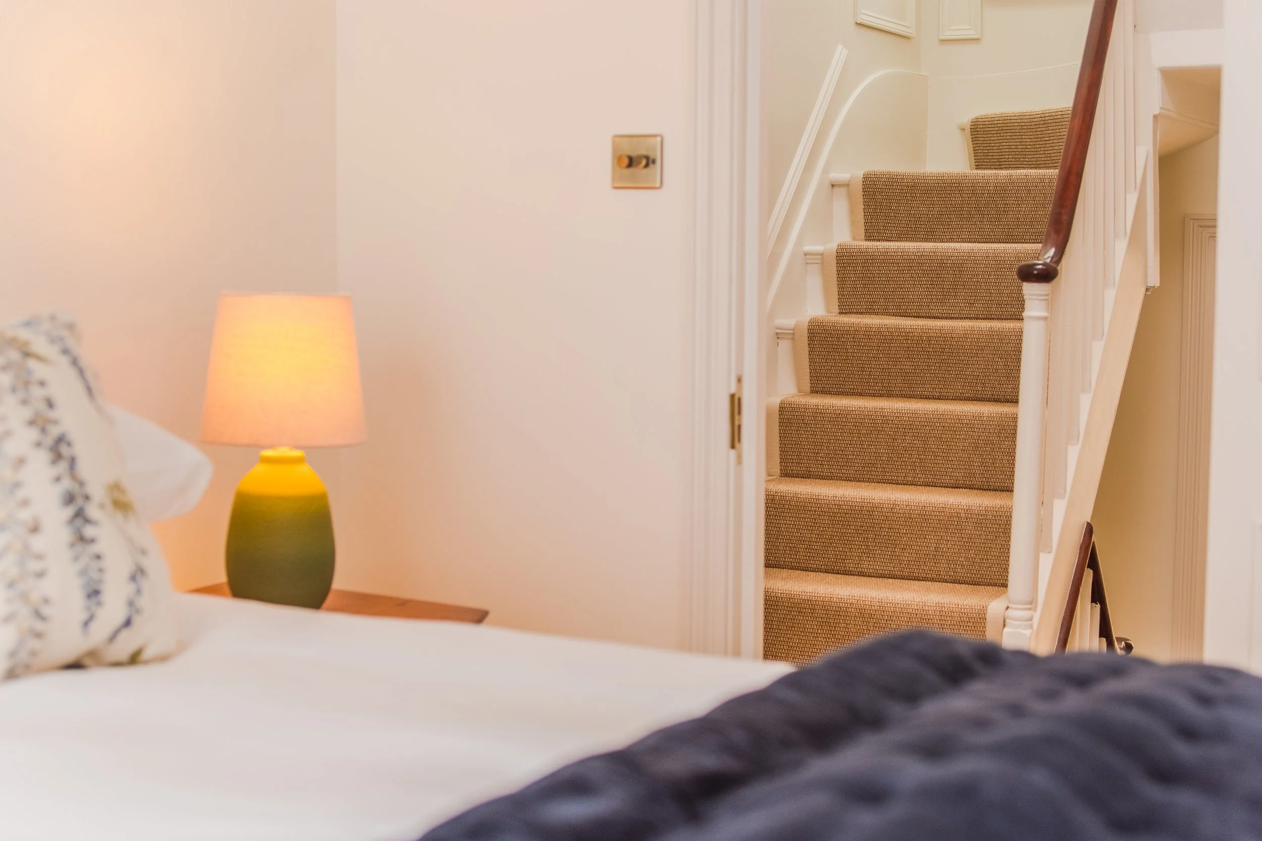 View of a bedroom with a bed, a nightstand with a lamp, and a staircase with beige carpeted steps and a wooden handrail.