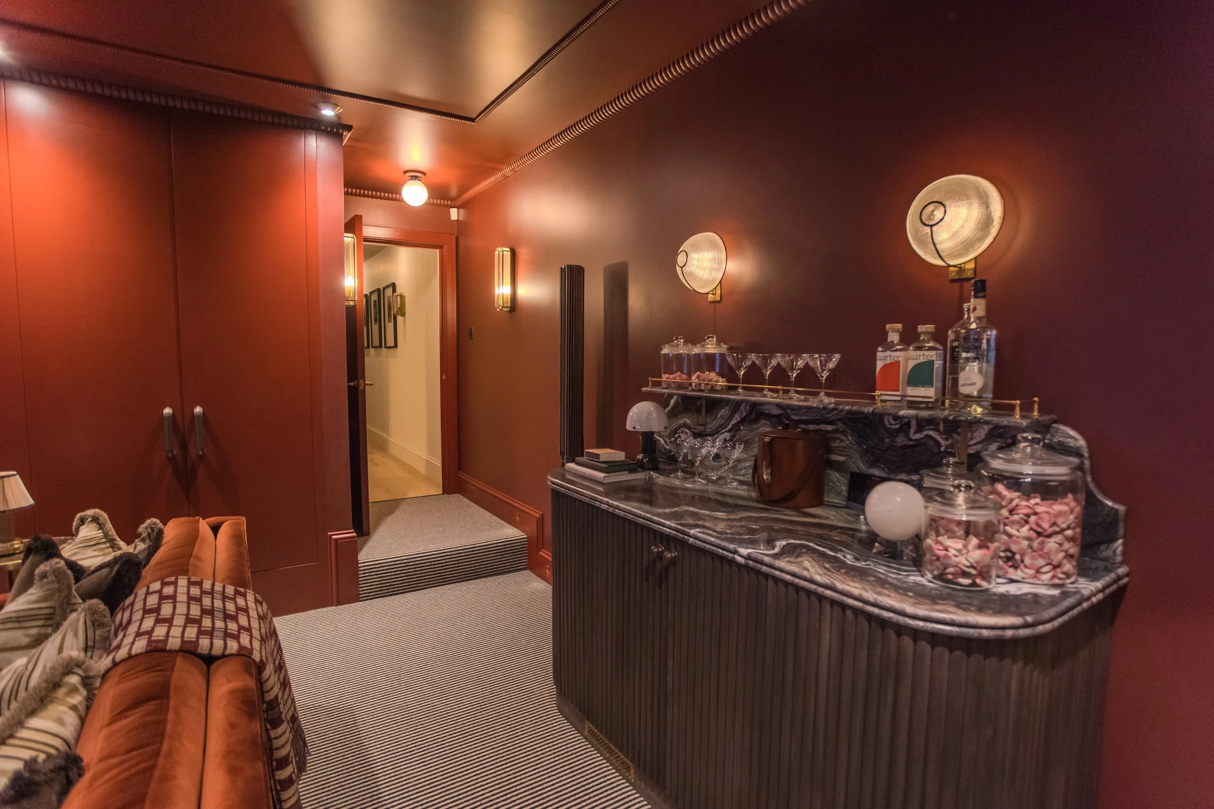 Jamie and Sophie Laing new house media room bar. A dimly lit, vintage-style living space with a red wall and a marble bar counter decorated with snacks, glasses, and bottles. There is a doorway leading to a hallway with framed artwork on the wall.