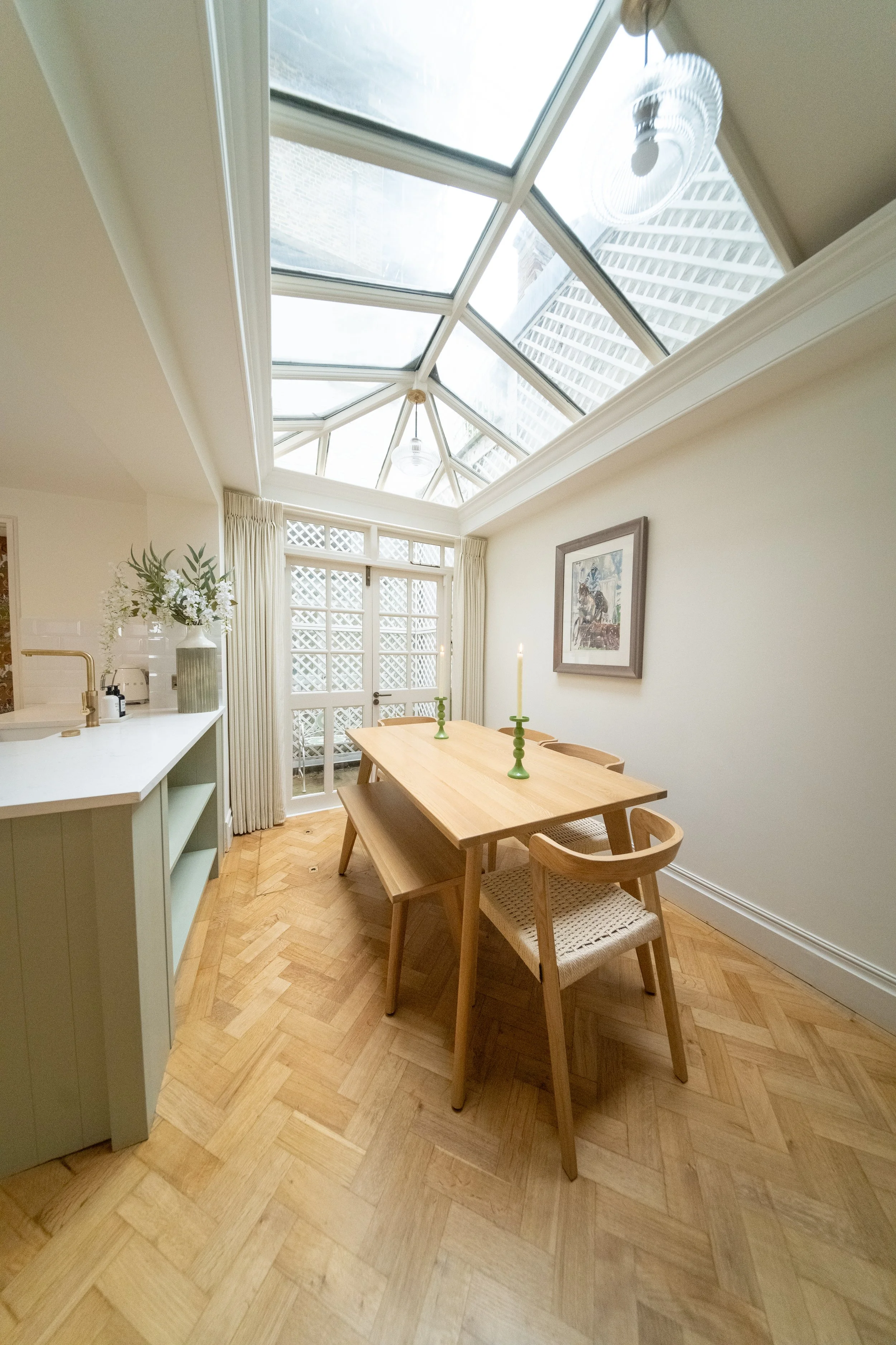 A sunlit dining area with a wooden table, three chairs, and two lit candles. A large glass ceiling and glass doors lead outside. A framed picture hangs on the wall, and a sideboard with plants and other decorative items is present.