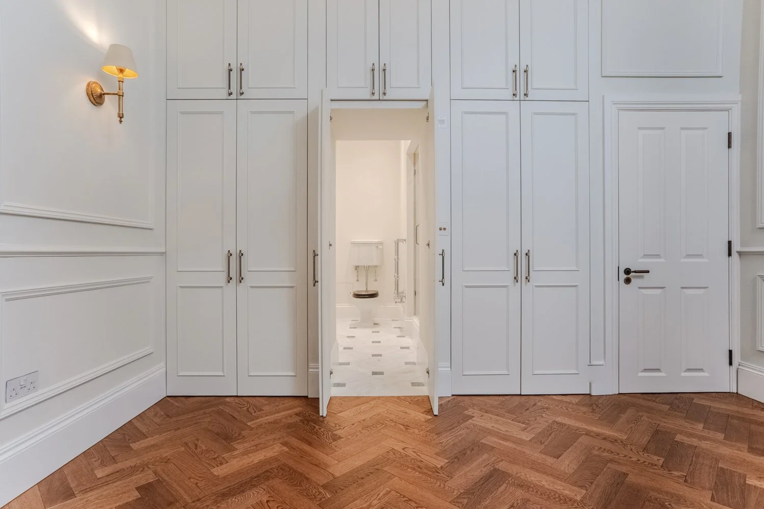A room with wood flooring, white paneled walls, and built-in white cabinets. An open door reveals a bathroom with a toilet and a window.