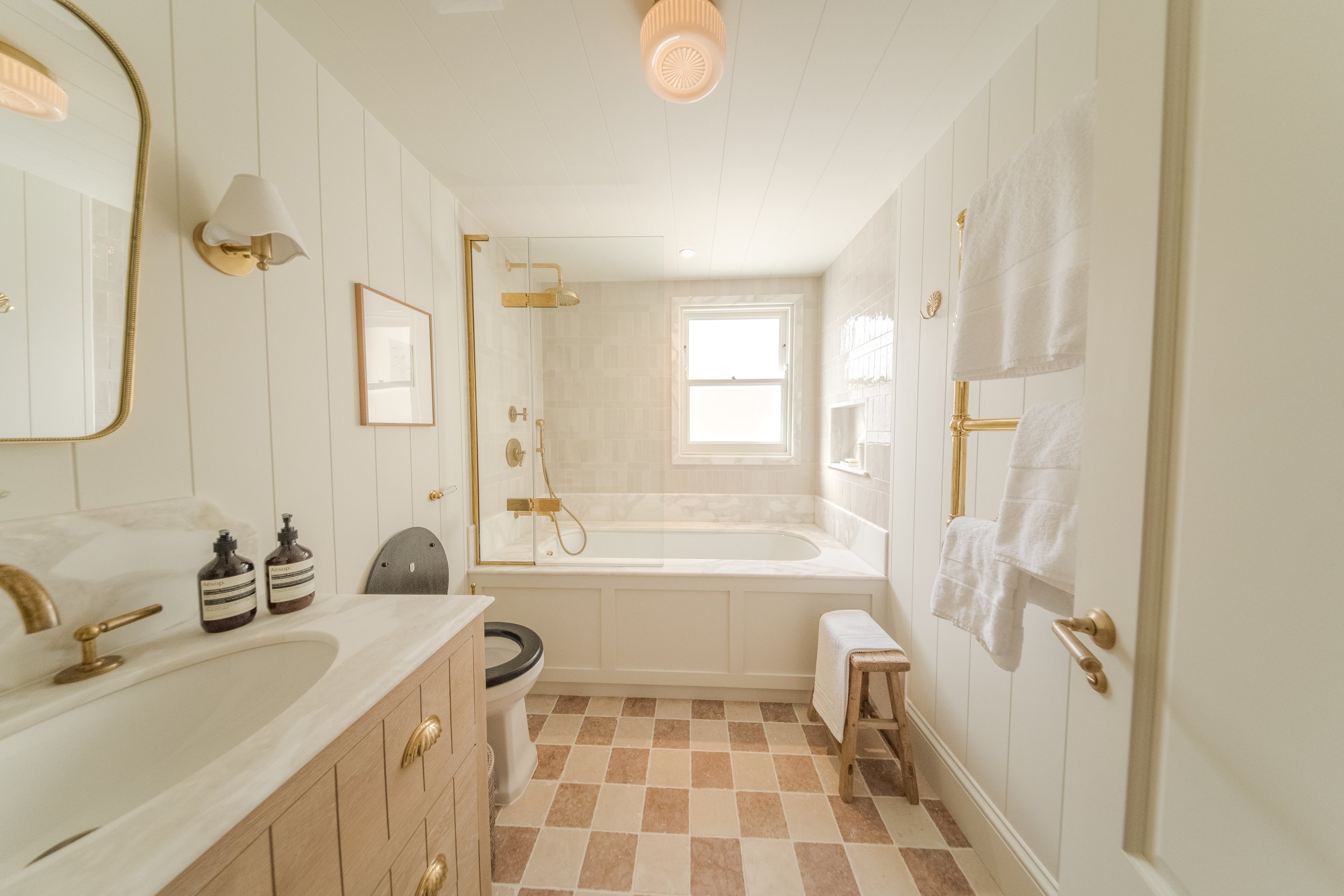 Jamie and Sophie Laing new home family bathroom. Bright bathroom with white panel walls, a bathtub with a window above, a glass shower door, a beige vanity with a marble countertop, and a towel rack with white towels.