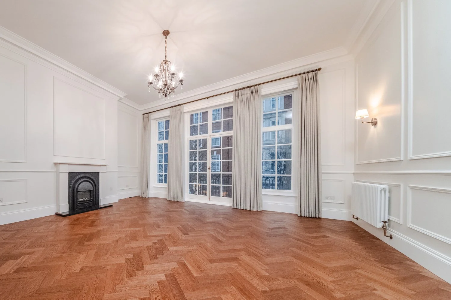 Empty living room with large windows, beige curtains, hardwood floors, white walls with decorative paneling, a fireplace, chandelier, wall sconce, and a radiator.