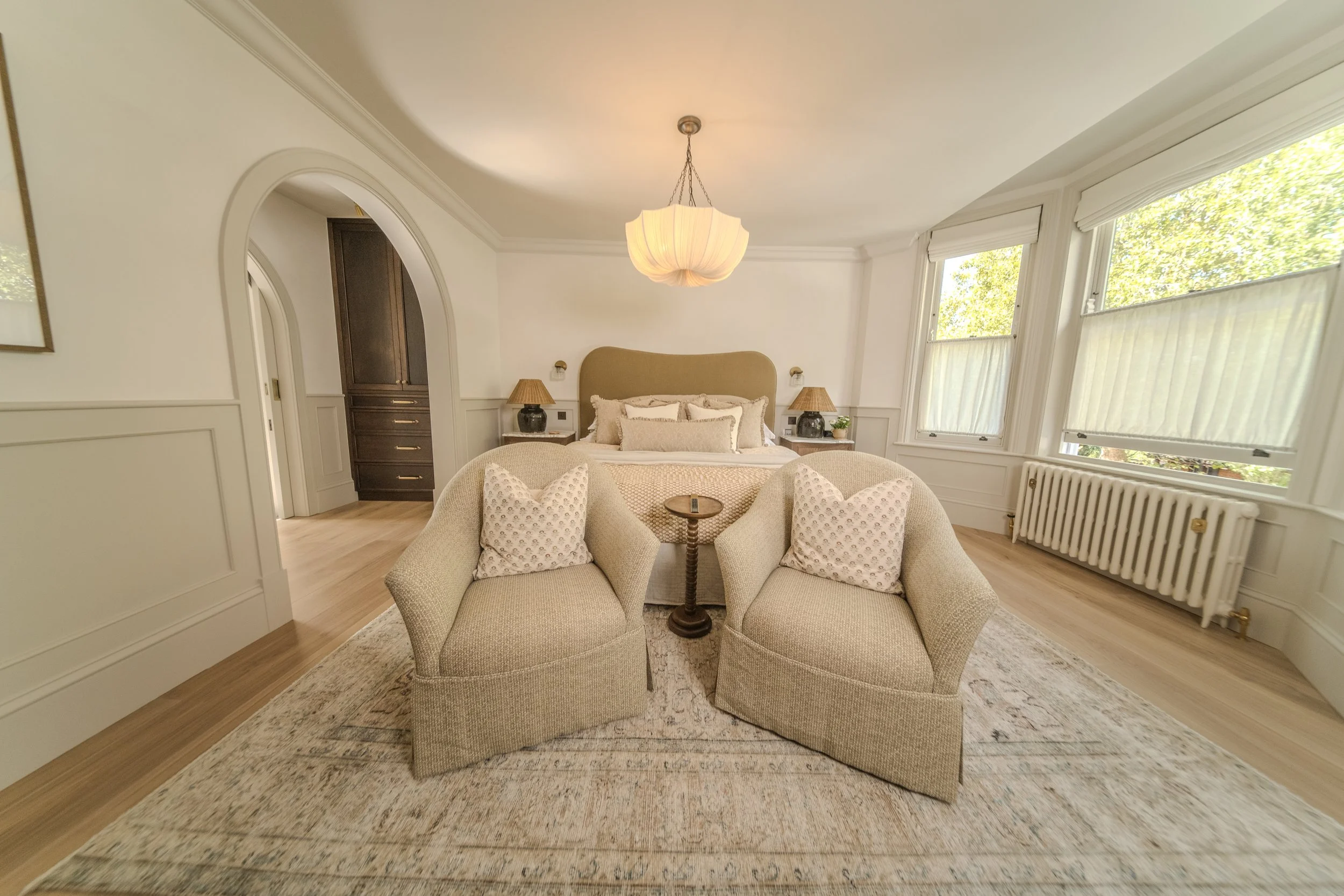 Bright bedroom with beige and white decor, featuring two beige armchairs with pillows, a bed with beige headboard, bedside lamps, large windows with blinds, and wooden flooring.