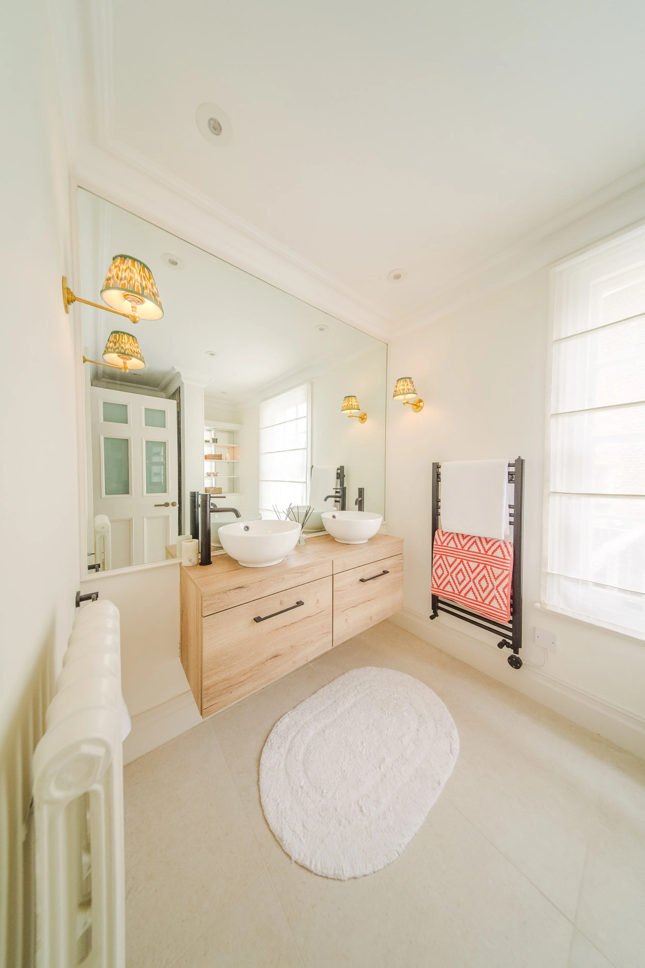 Bathroom with a large wall mirror, two vessel sinks on a wooden vanity, wall-mounted lamps, a window with blinds, a towel rack with a decorative towel, a white rug, and a radiator.