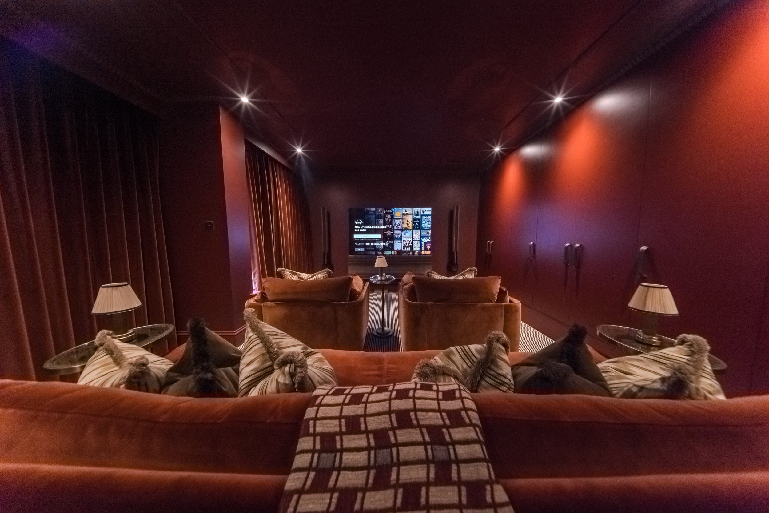 A luxurious home theater room with a large flat-screen TV, two brown armchairs, a plush reddish-brown sofa with striped pillows, side tables with lamps, dark red curtains, and dim lighting.