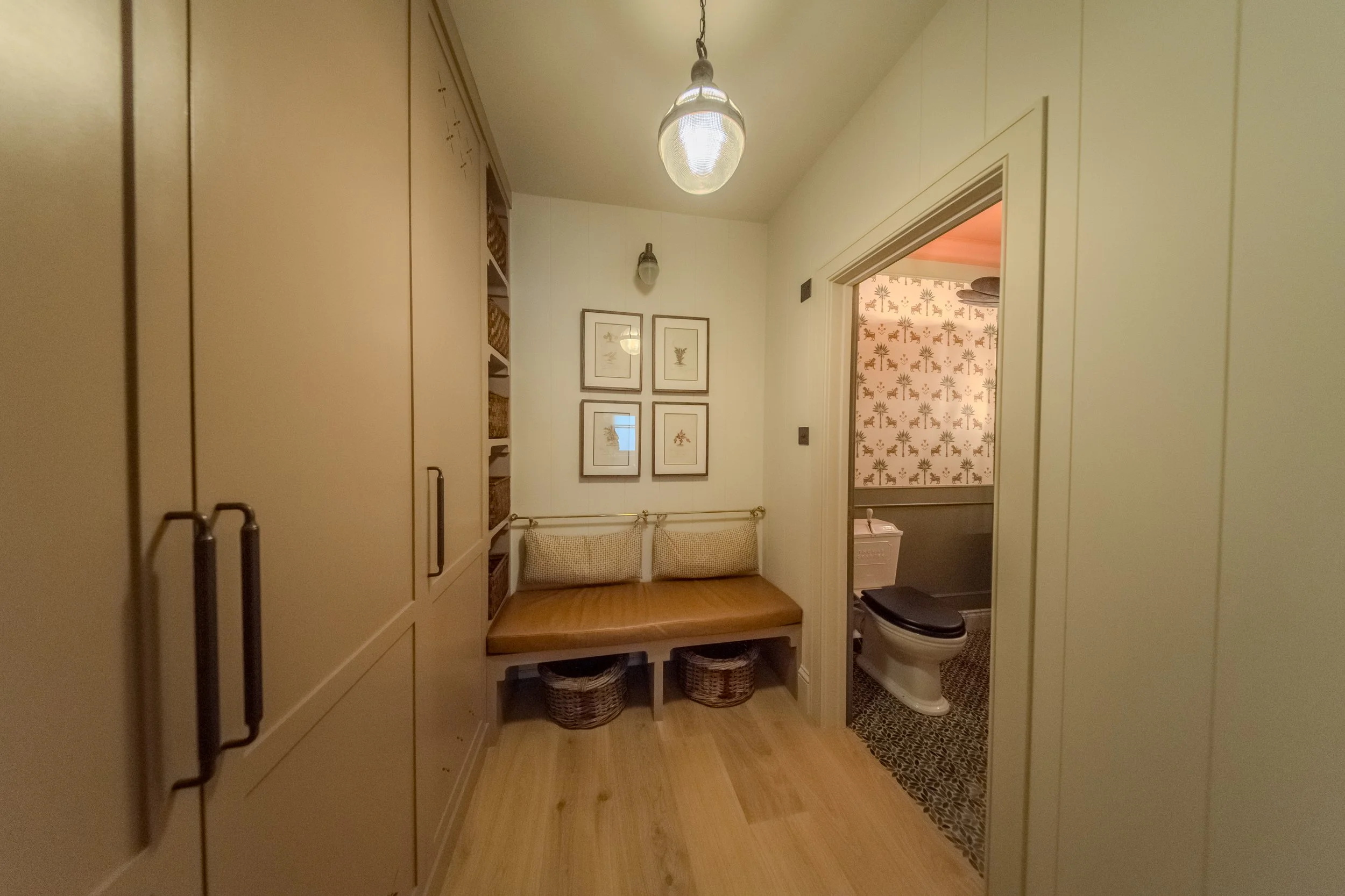 Hallway with built-in beige cabinetry, a bench with pillows, framed botanical prints on the wall, and a view into a bathroom with wallpaper featuring palm trees and a toilet.