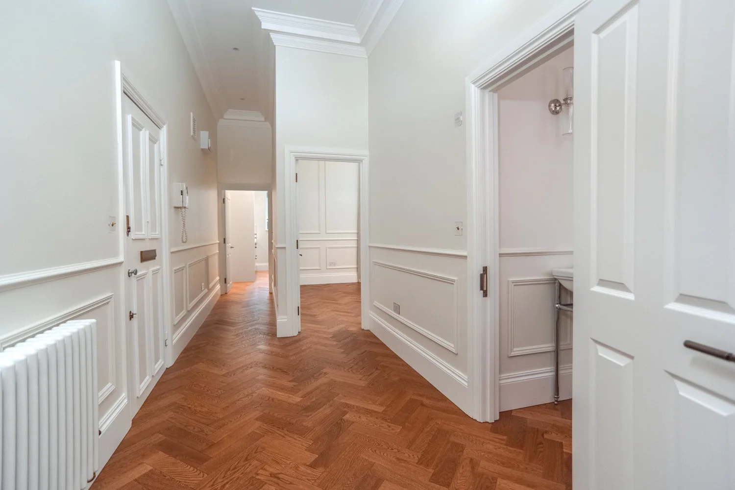 Empty hallway with white walls, intricate crown molding, and polished wooden herringbone floors, leading to several open doors.