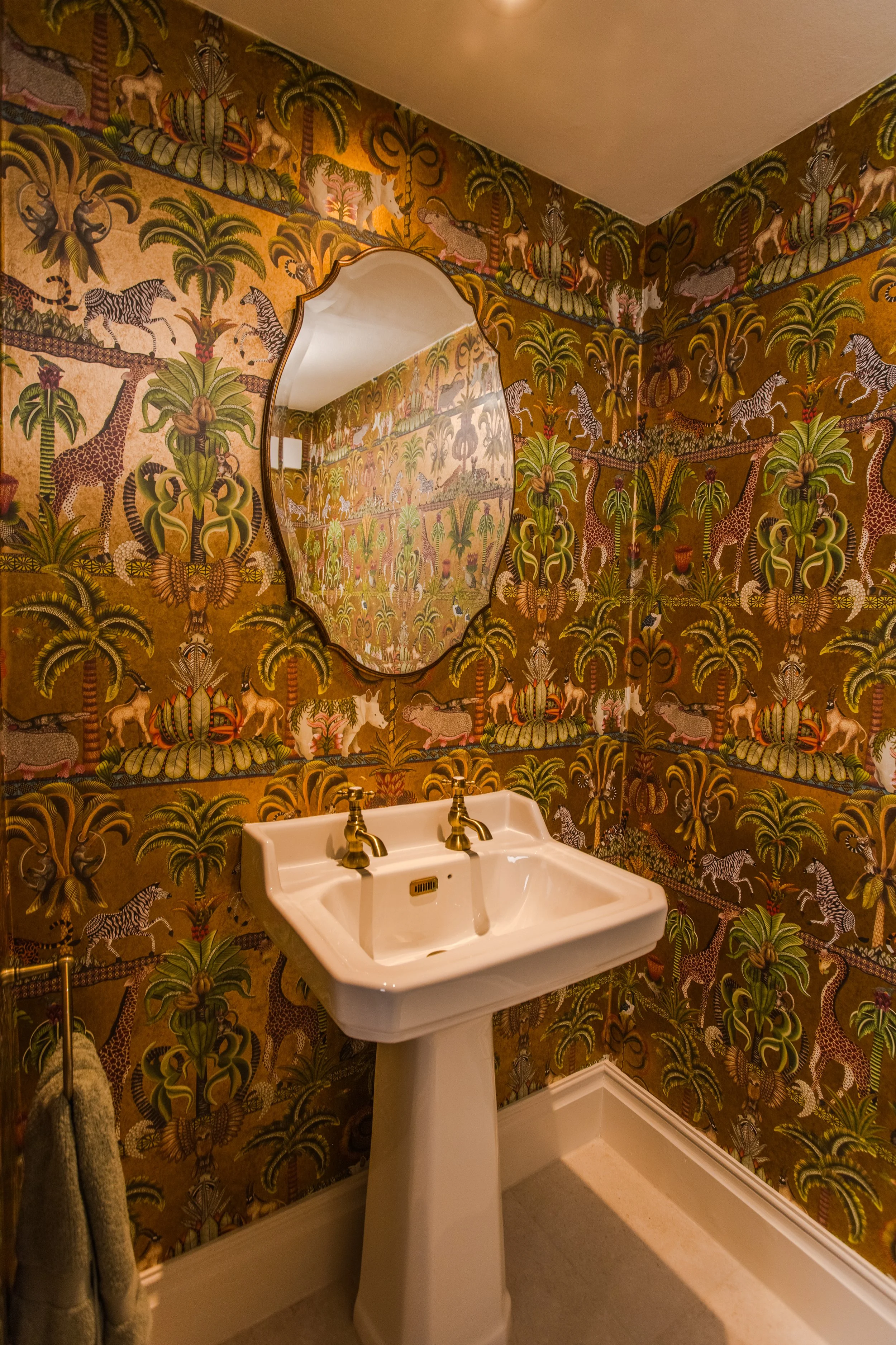 Bathroom with jungle-themed wallpaper featuring animals like giraffes and zebras, a white pedestal sink with two brass faucets, and a mirror reflecting the wallpaper.