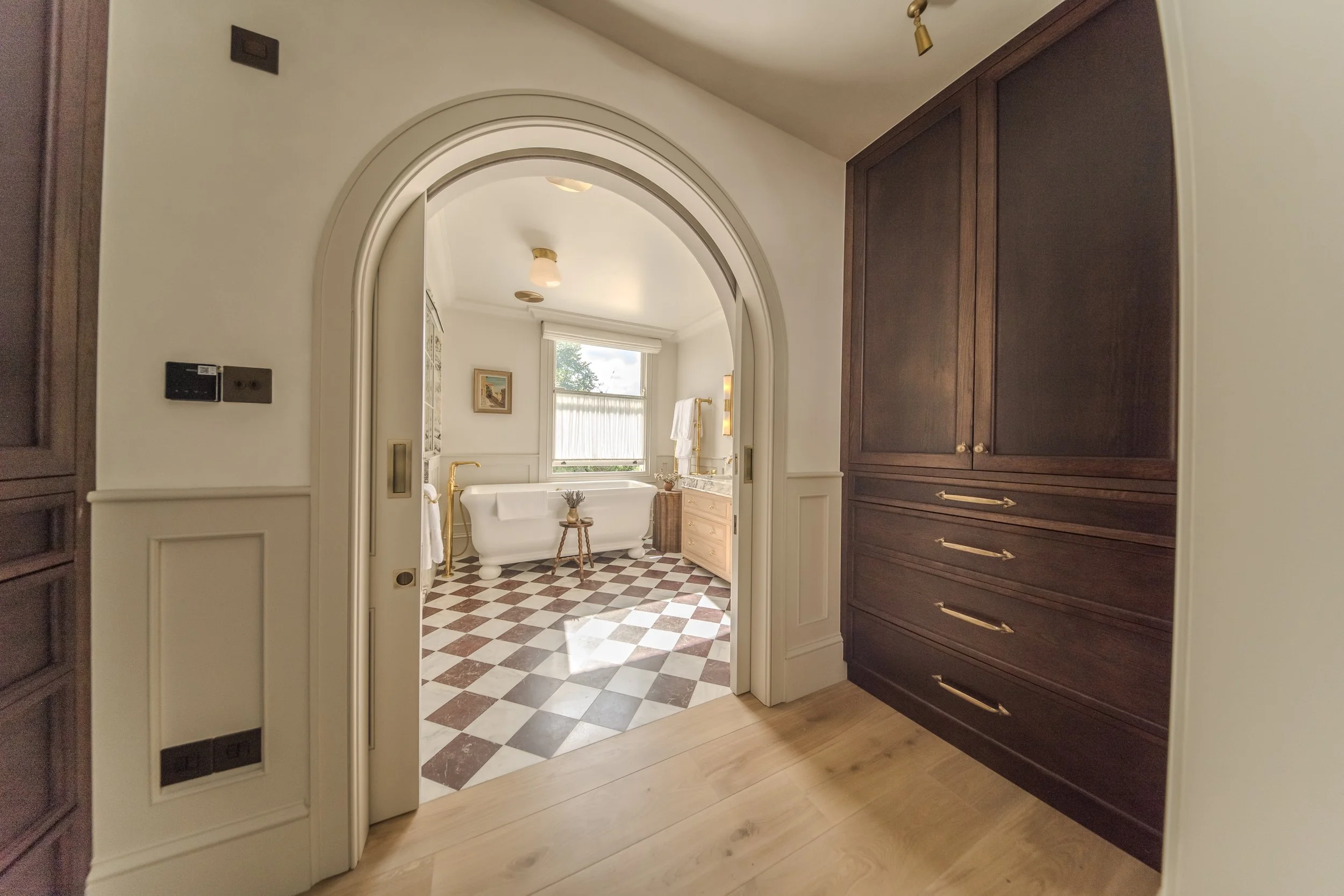 Jamie and Sophie Laing new house joinery and bathroom arch. Bathroom with bathtub, window, and checkered tile floor, viewed through an arched doorway from a room with beige walls and dark wooden cabinetry.