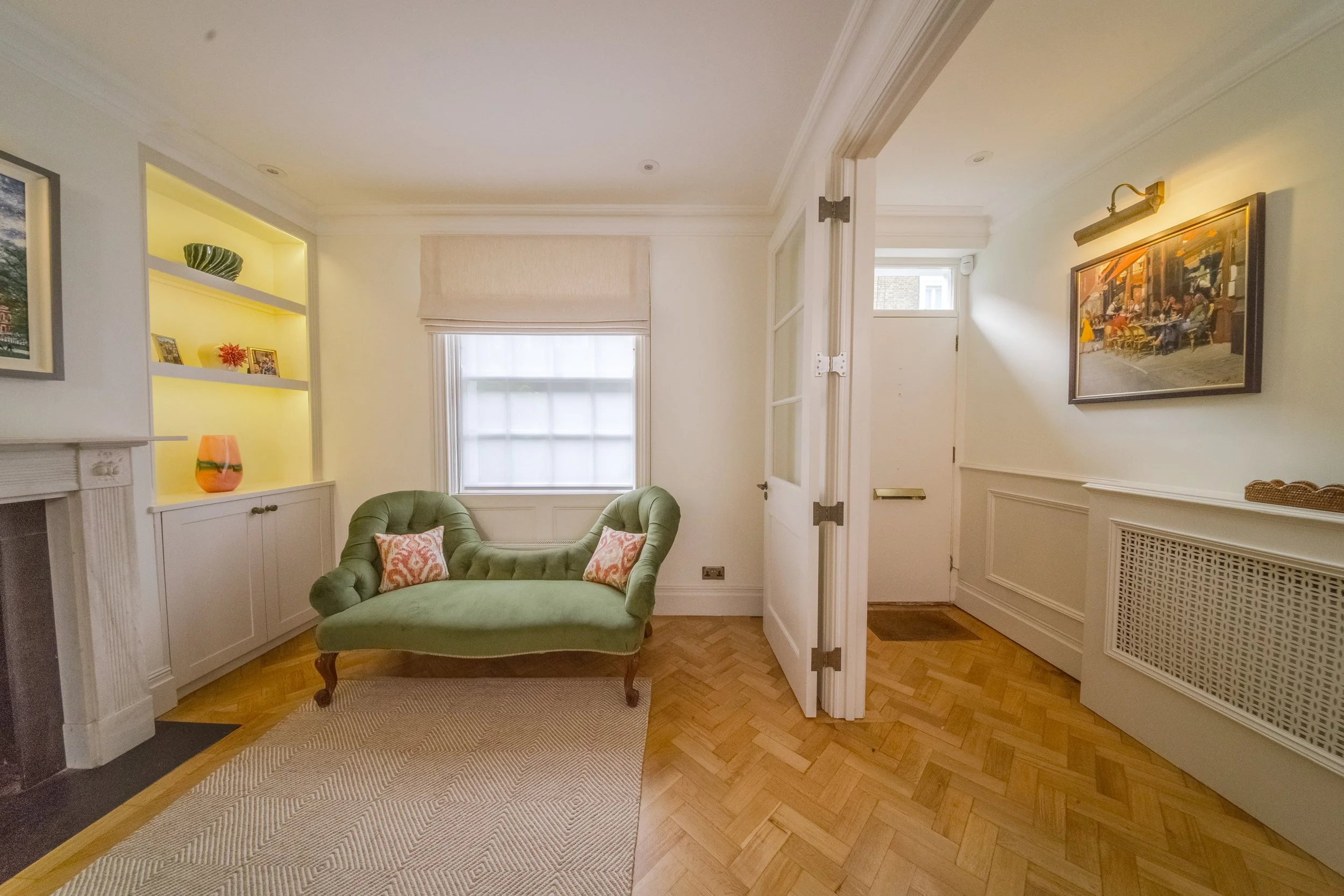 Living room with a green vintage loveseat, window with blinds, built-in bookshelf with decorative items, artwork on the wall, and a doorway leading to an entryway.