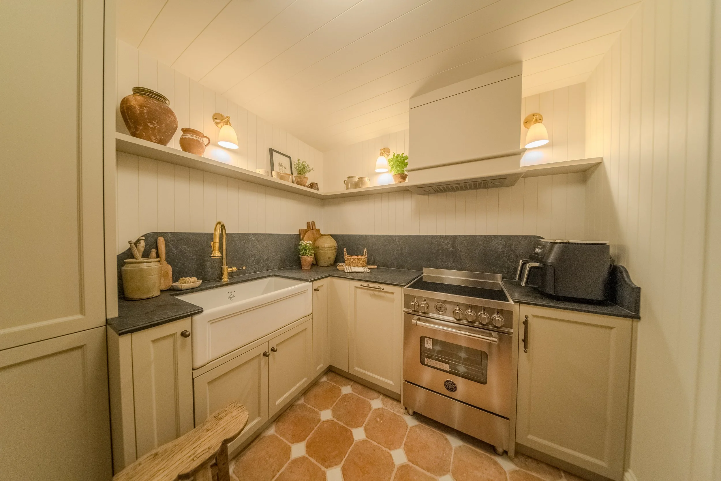 Jamie and Sophie Laing new home utility room. A cozy kitchen and utility room with cream-colored cabinets, black countertops, and a white farmhouse sink. Open shelving above displays decorative pottery and small plants. 