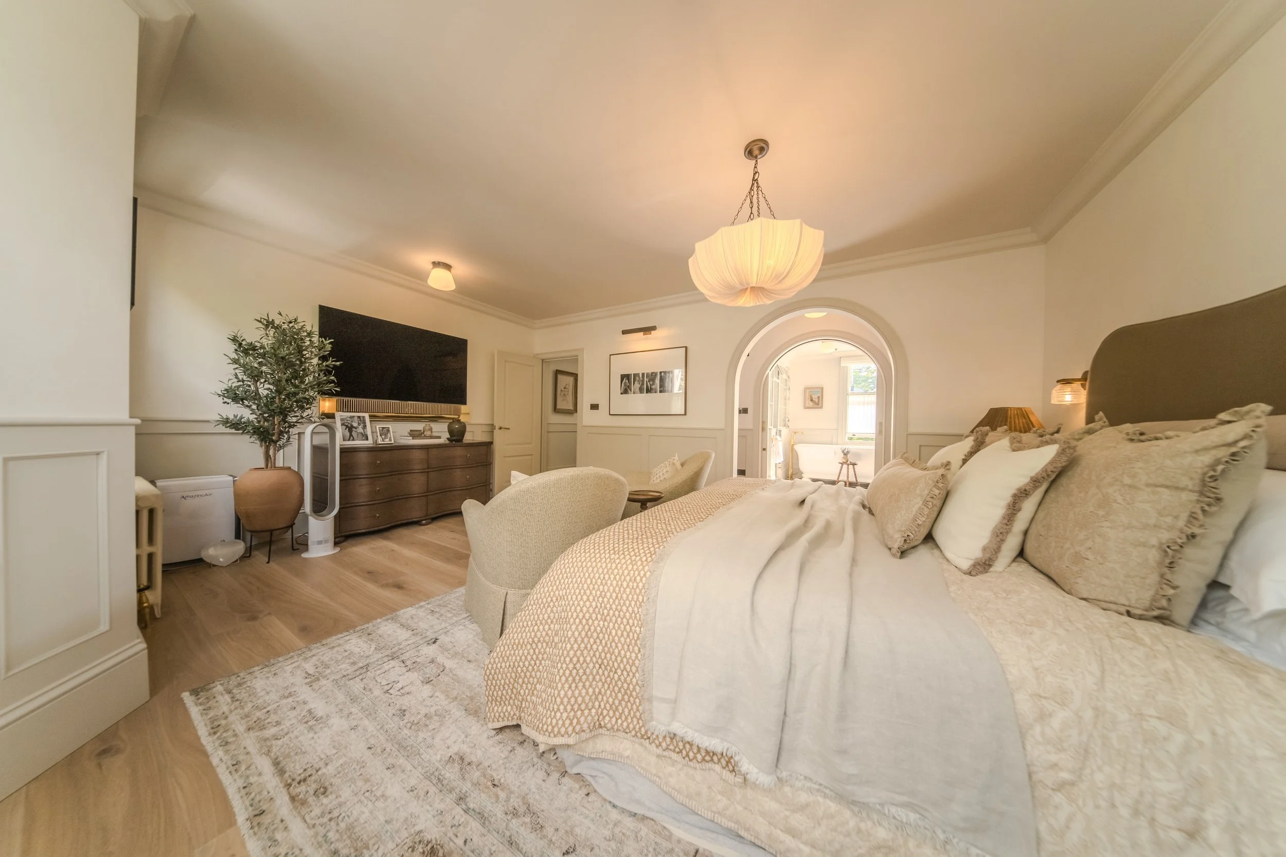 Jamie and Sophie Laing renovated bedroom with a large bed, beige bedding and multiple pillows. Light-colored furniture, a chandelier, and an arched doorway leading to another room with natural light. Designed by House 9 design 