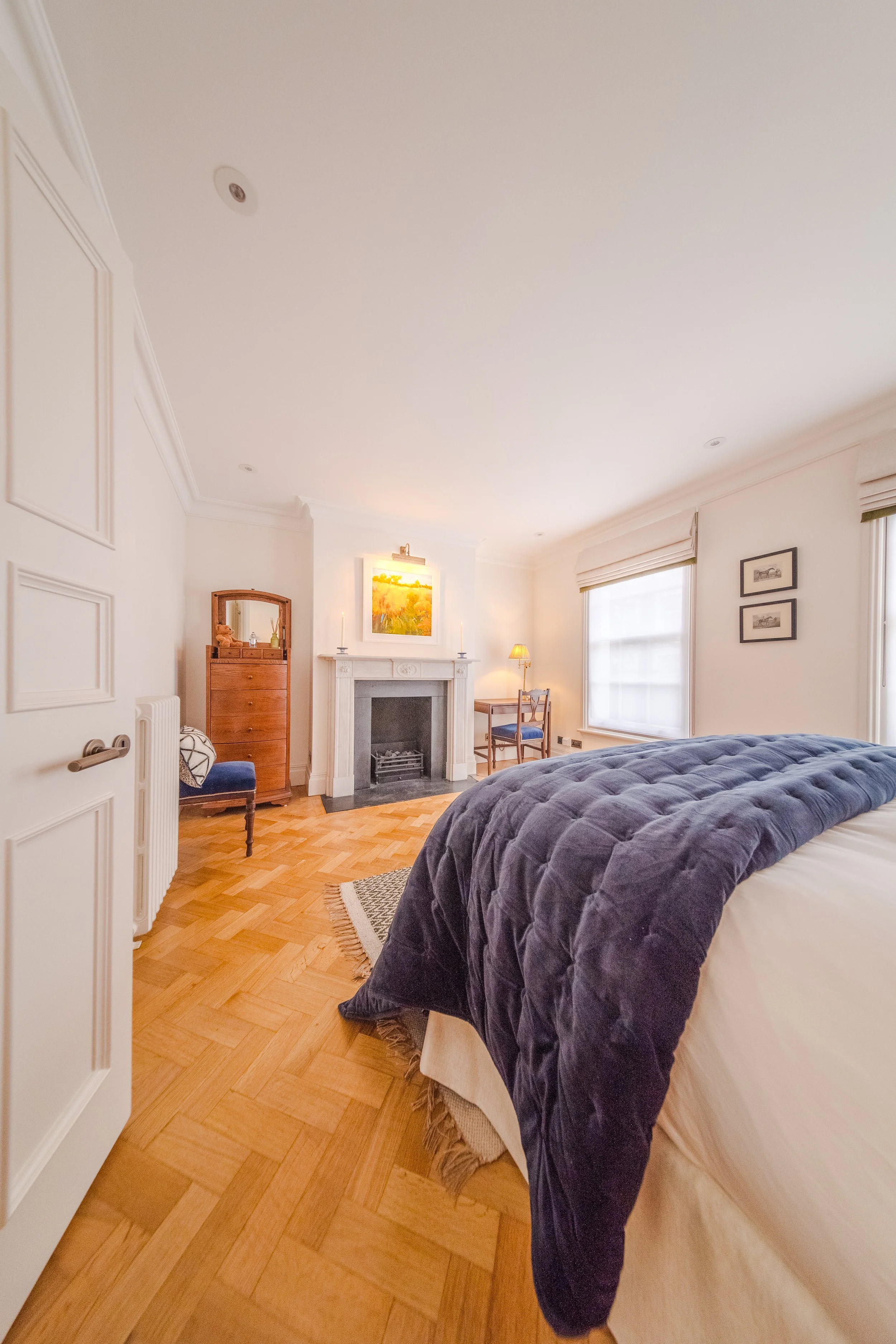 A cozy bedroom with a fireplace, wooden floors, white walls, large windows with blinds, a bed with a navy blue quilt, and framed artwork on the wall.