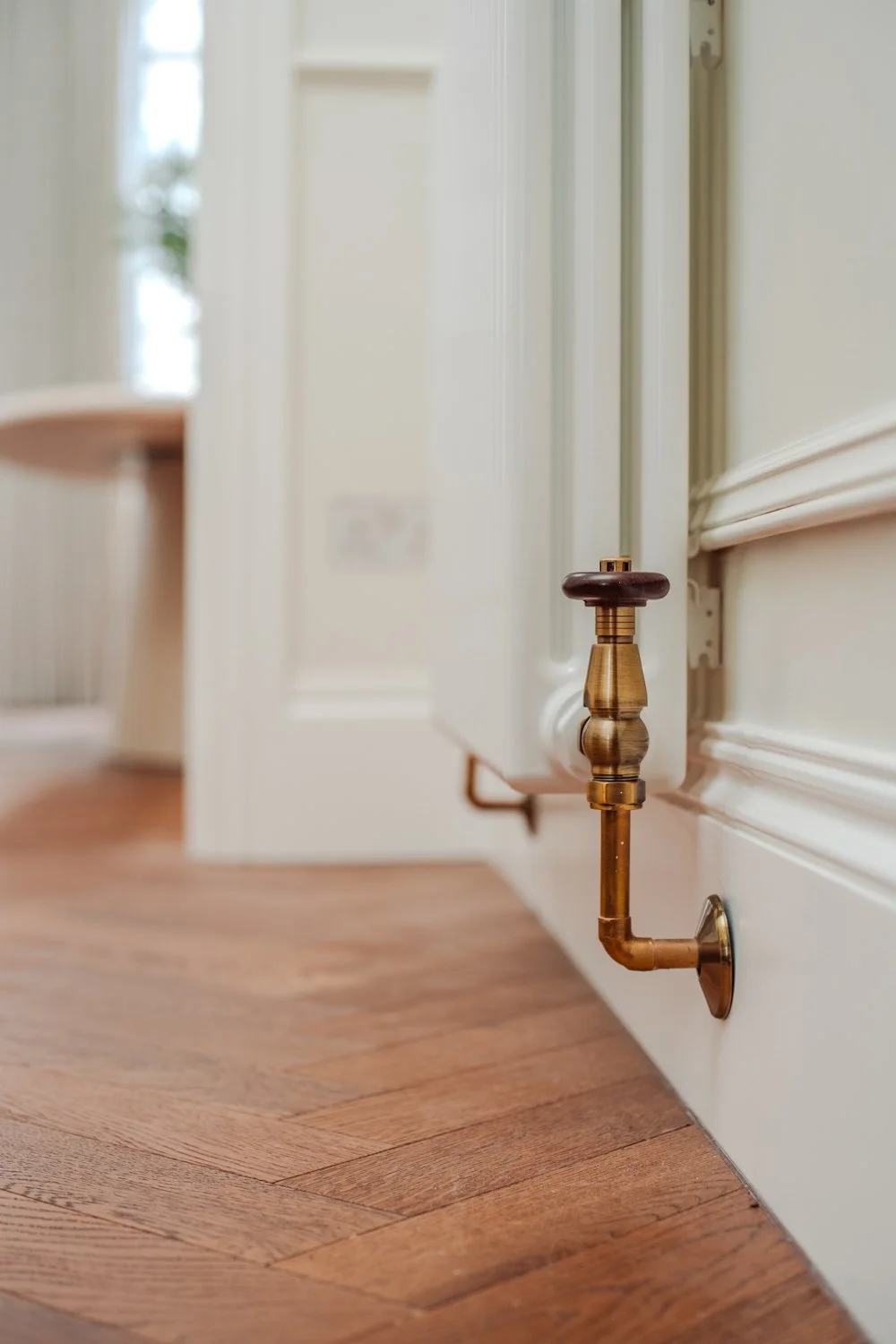 Close-up of a copper pipe with a valve, attached to a white radiator in a room with wooden flooring and a door in the background.