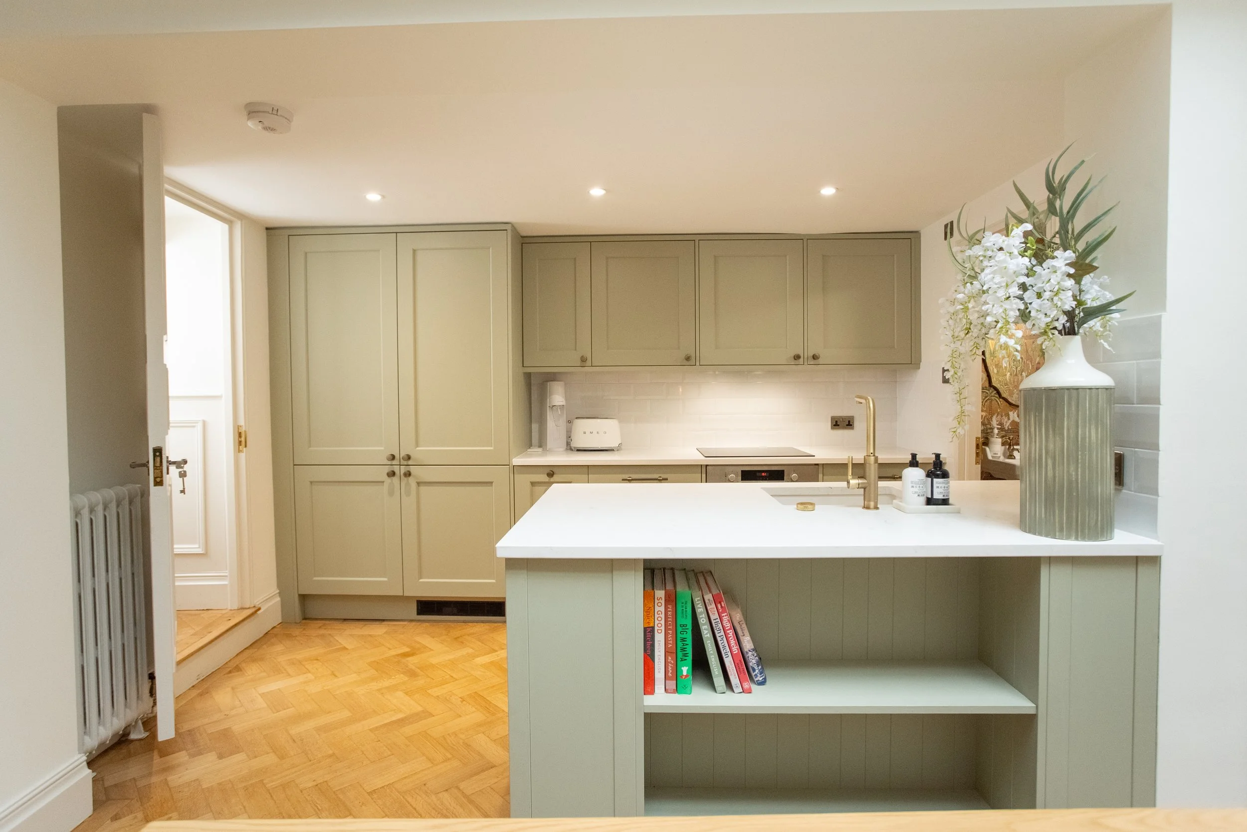 Modern kitchen with light beige cabinets, white countertop, and gold faucet. Decor includes a large vase with white flowers and leafy greenery, with a few books on the open shelf of the kitchen island.