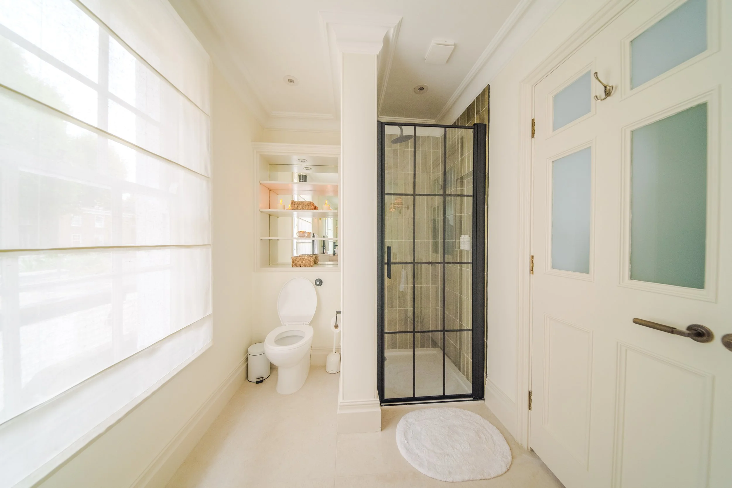 A bright bathroom with a window covered by a white shade, a toilet, open shelves with baskets and toiletries, a glass shower with black framing, and a white door with frosted glass panels.