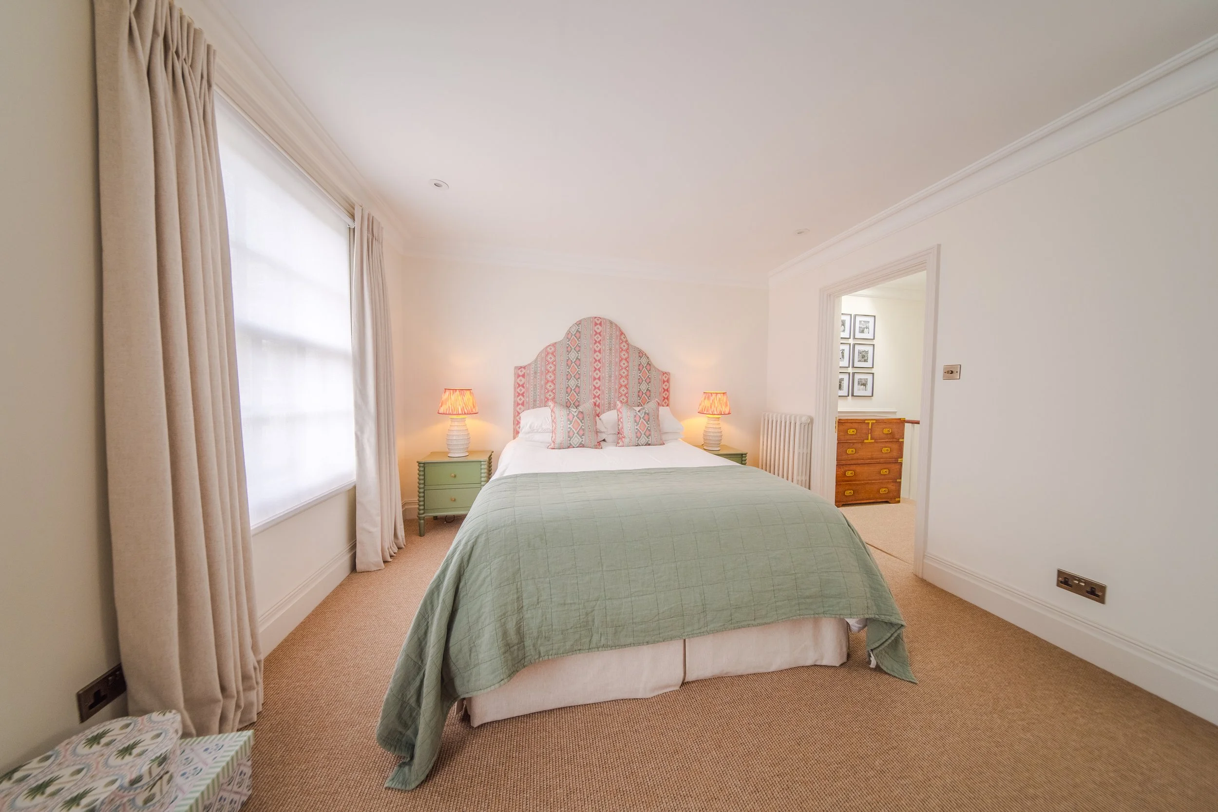 Bedroom with bed, green quilt, pink patterned headboard, two matching pink lamps, teal nightstands, beige carpet, window with beige curtains.