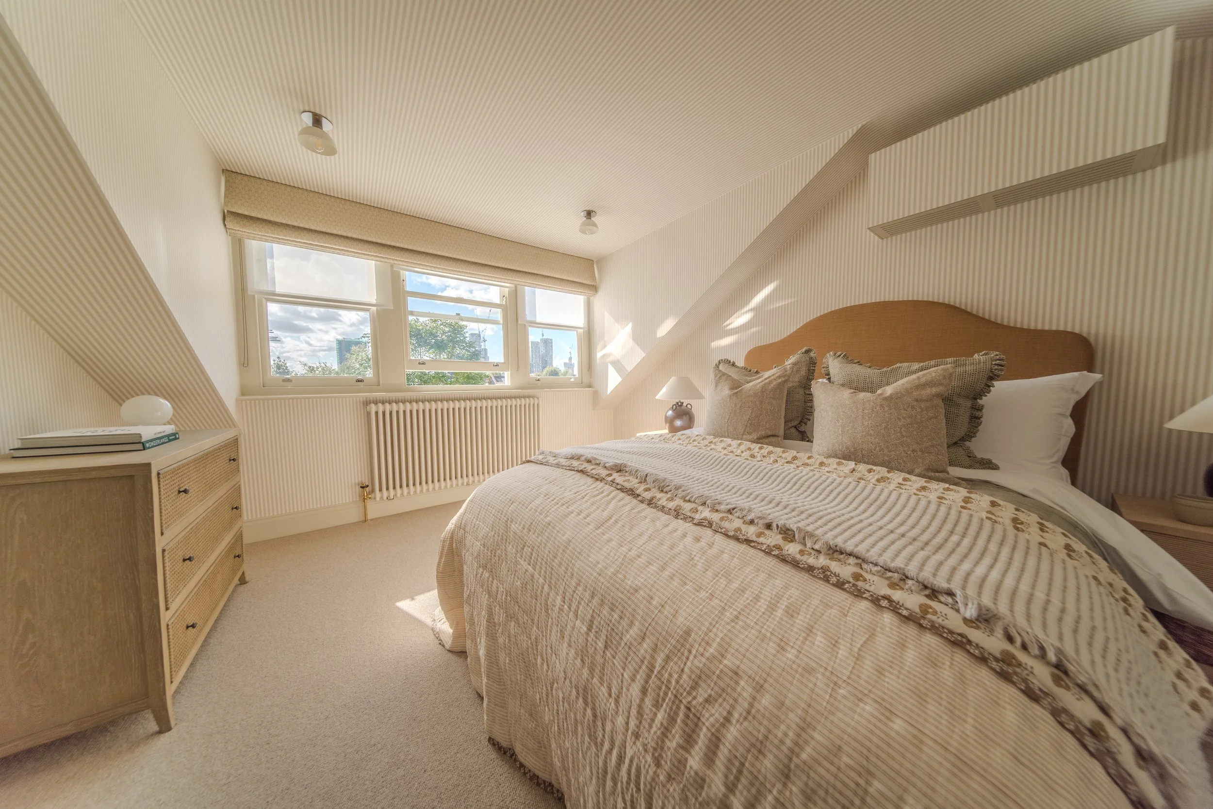A cozy bedroom with a large bed, decorative pillows, a wooden headboard, nightstands with lamps, a dresser, and a window with city skyline view outside.
