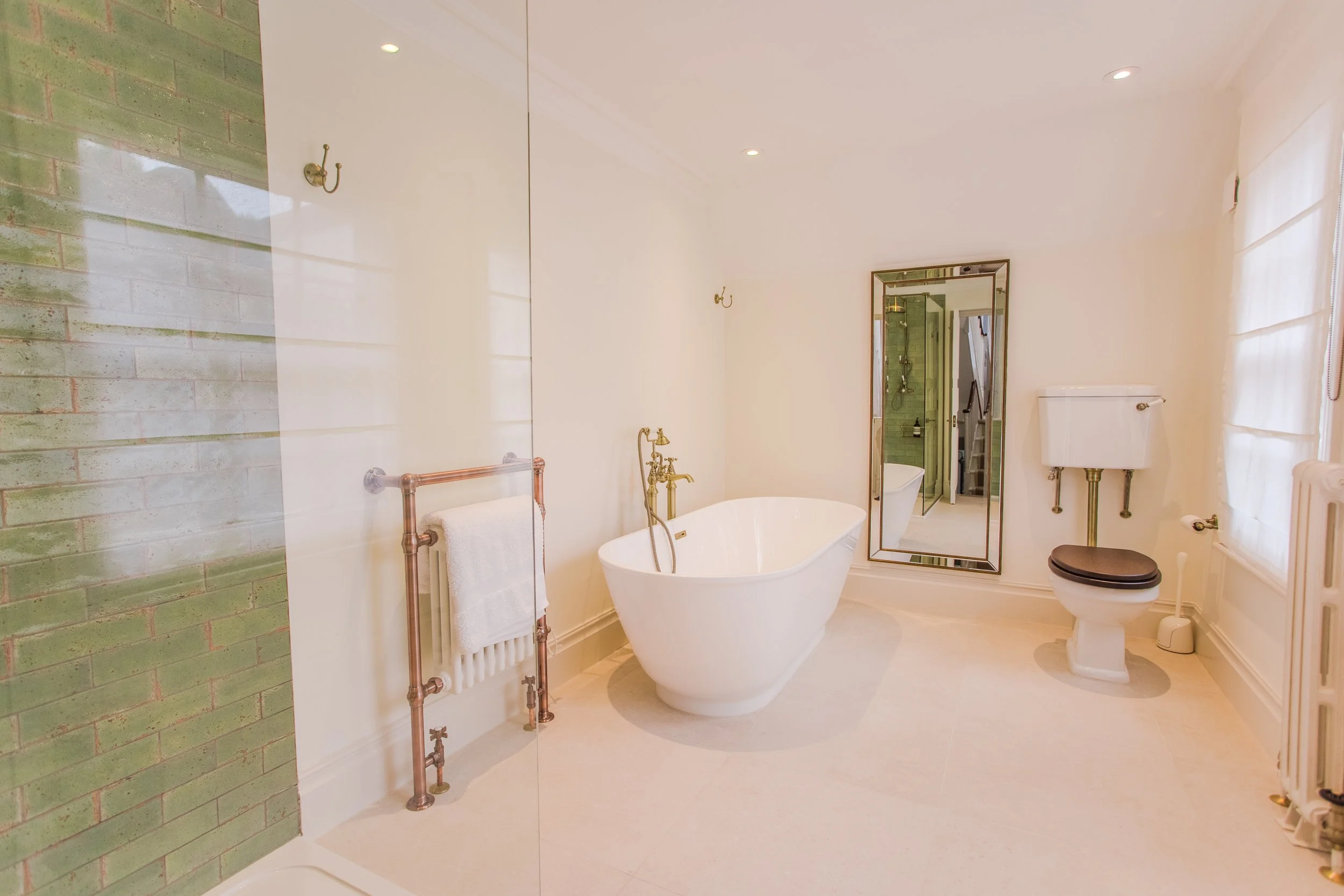 Bright bathroom with a freestanding bathtub, toilet with black seat, mirror, towel rack, and green tiled shower area.