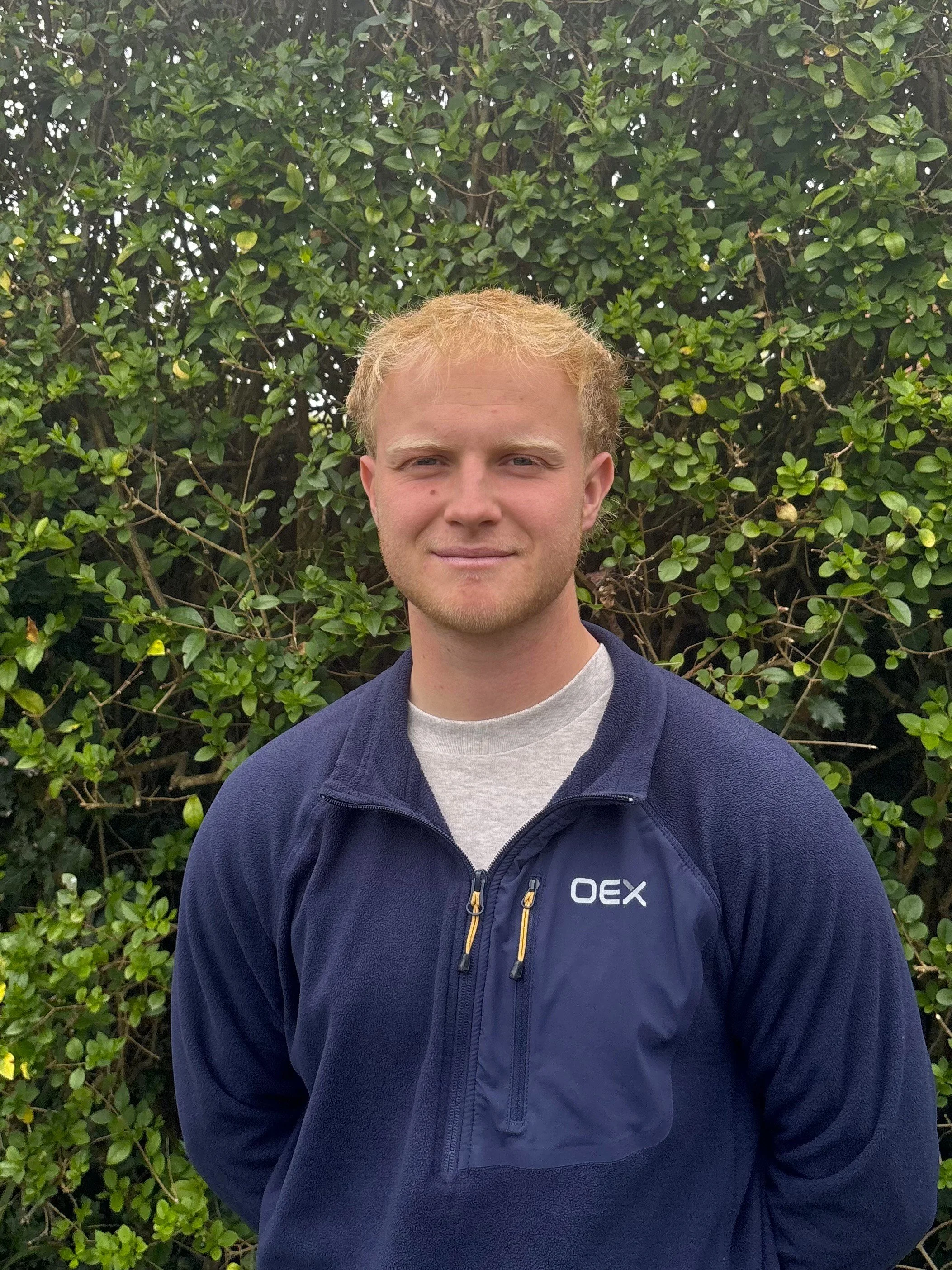 A young man of fair skin and short blonde hair , standing in front of a green hedge, wearing a navy blue jacket and grey tshirt