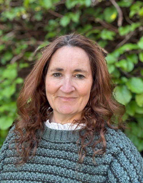 A woman with long, wavy brown hair wearing a gray sweater with a white ruffled shirt underneath, standing outdoors in front of green foliage.