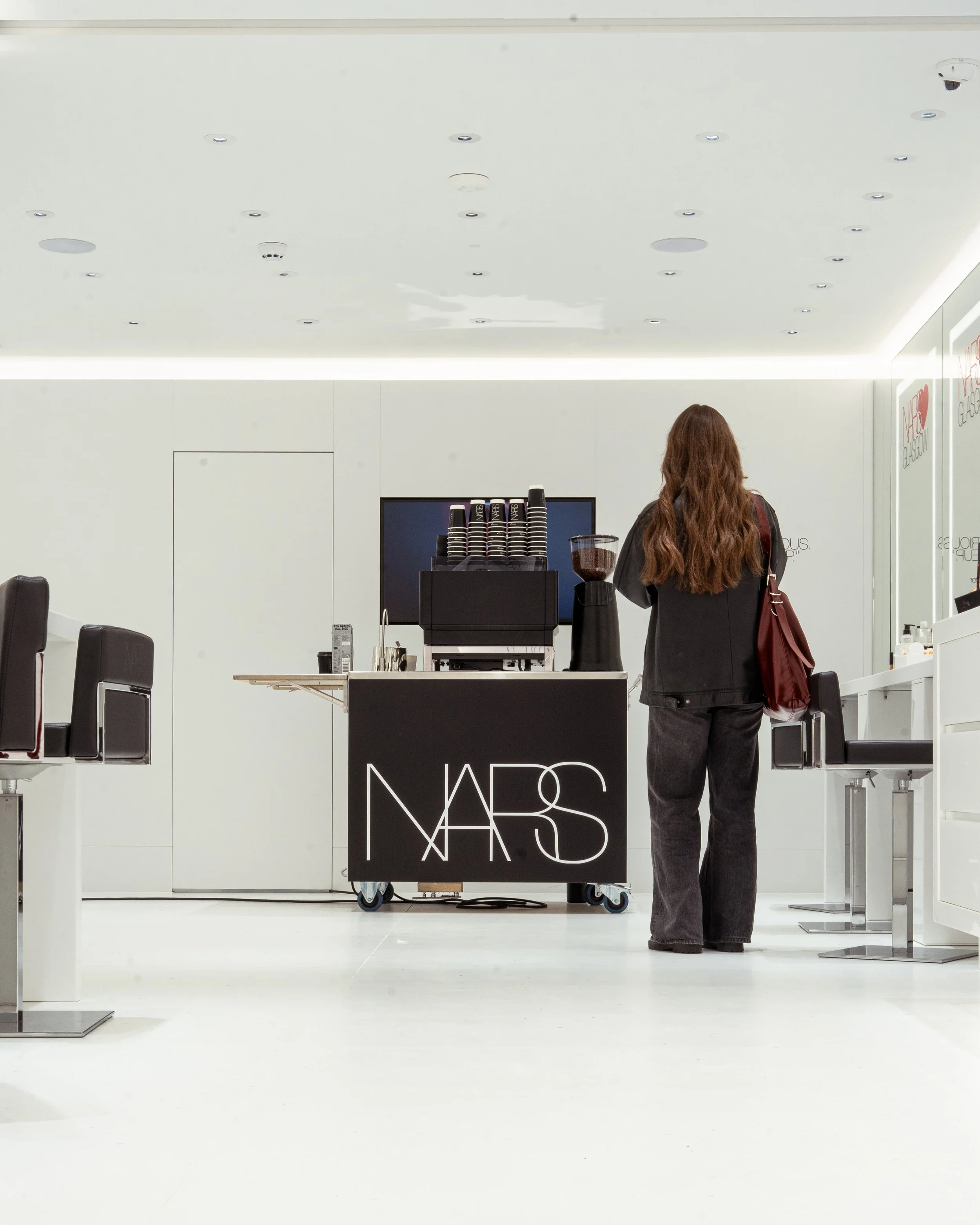 A woman with long brown hair, dressed in black, standing at a NARS cosmetics counter inside a minimalist, white store.