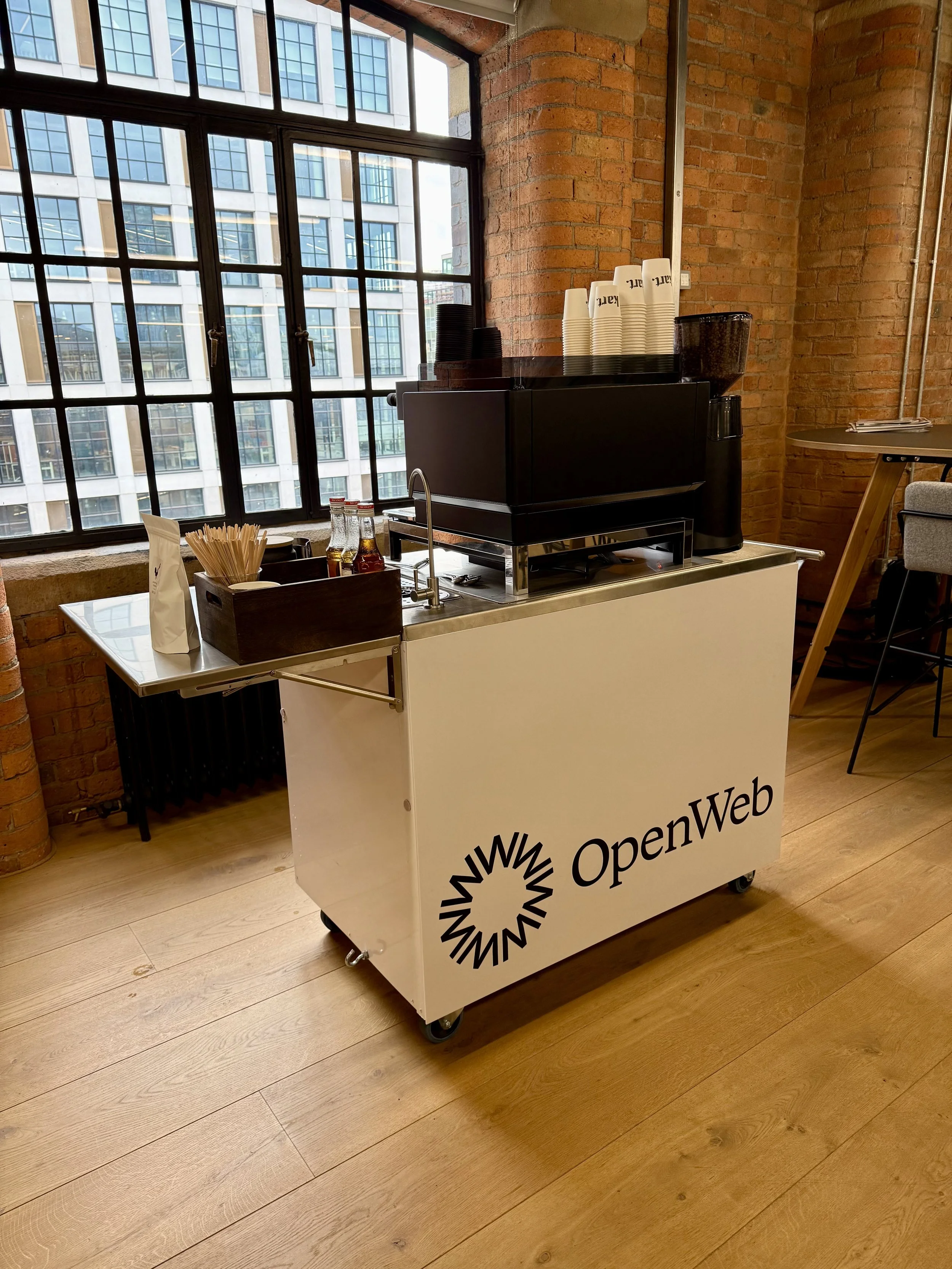 A mobile coffee station with a black coffee machine, cups, stir sticks, and bottles of syrup in a room with large windows, brick walls, and wooden floors, branded with the OpenWeb logo.