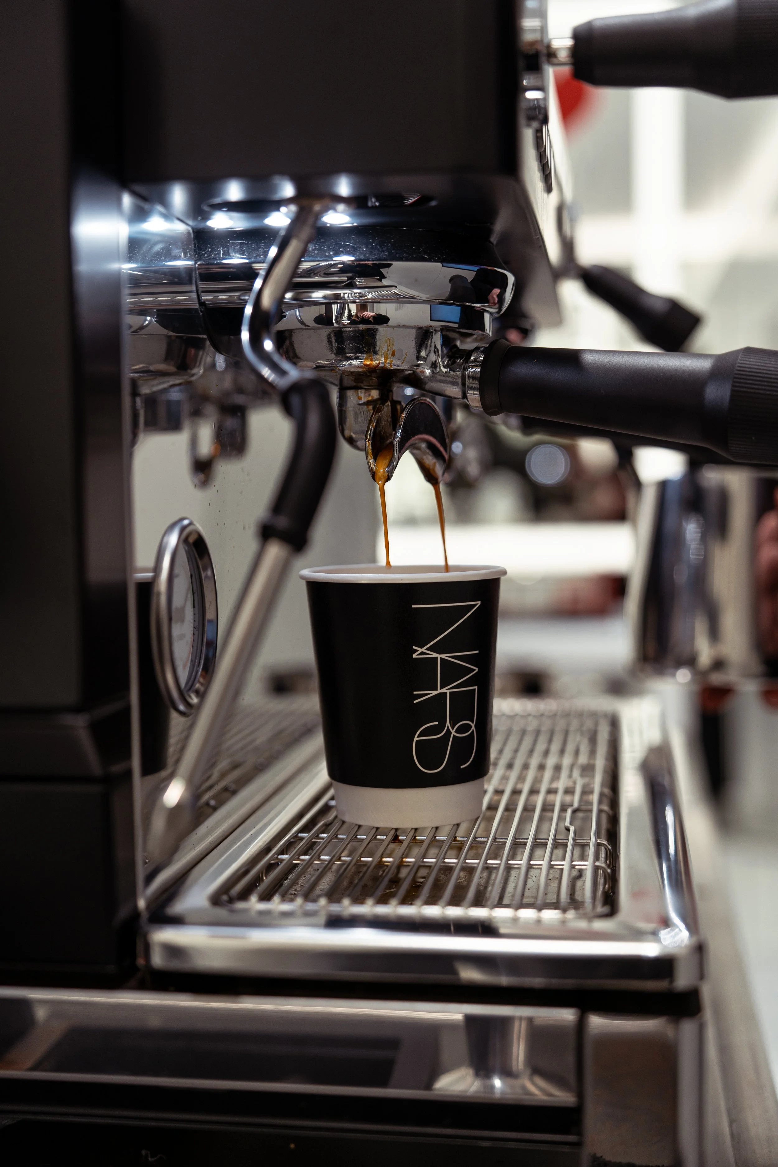 Close-up of a coffee machine pouring espresso into a black coffee cup with white inside, labeled 'NARS' in white. The espresso flows into the cup on a metal drip tray.