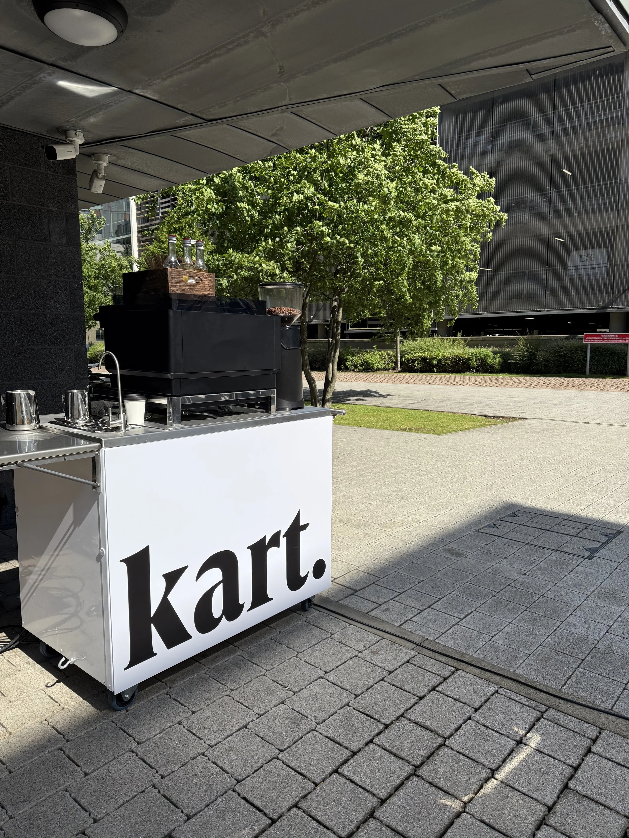 A portable coffee cart labeled 'kart' in an outdoor urban setting with trees, buildings, and a paved area.