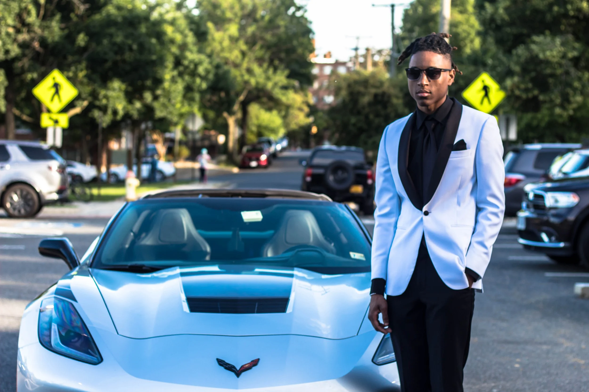 A man in a white blazer, black shirt, and sunglasses standing next to a silver sports car in a parking lot with trees and other cars in the background.