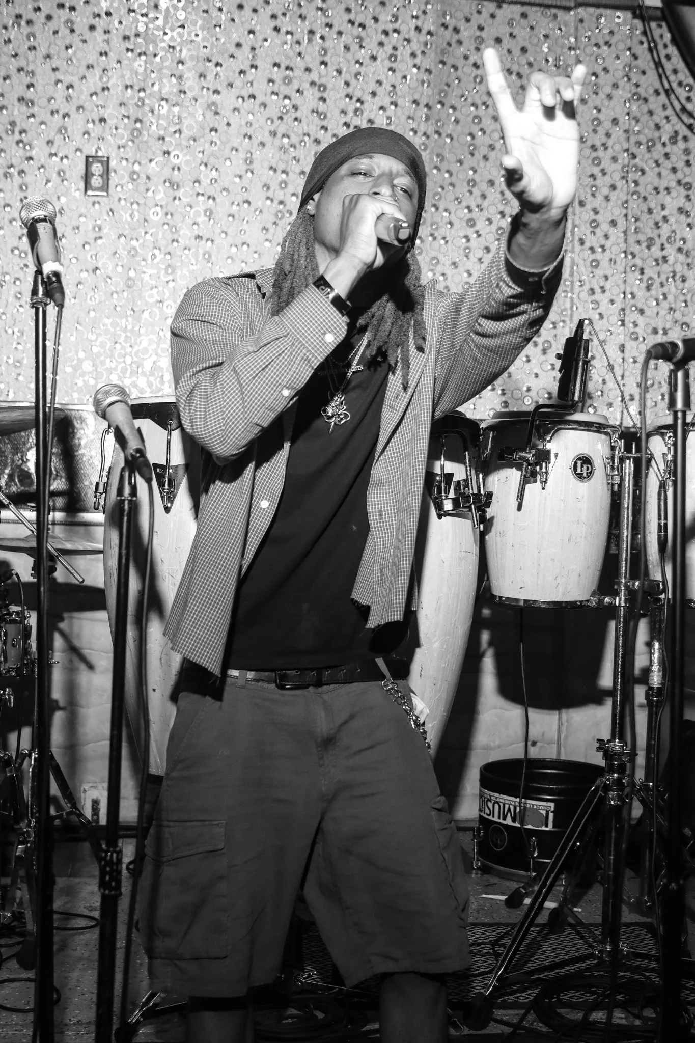 A male singer with dreadlocks, wearing a beanie and casual clothing, performing on stage with a microphone in hand. There are drums and other musical equipment visible in the background.
